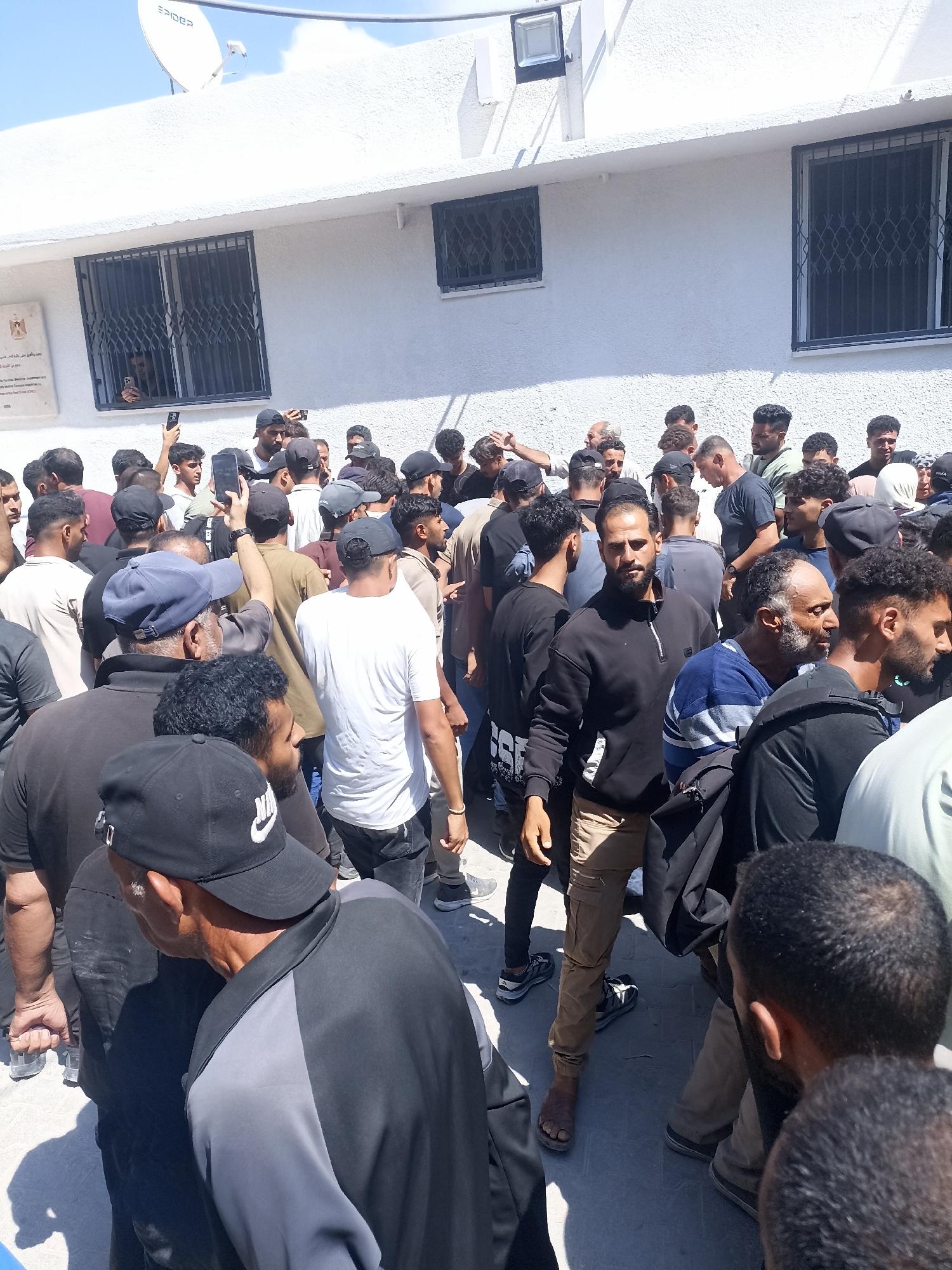 A group of men are gathered outside a white building, some holding cell phones. The building has windows with barred coverings. One window has a person looking out of it. A satellite dish is mounted on the roof of the building. The ground is paved with grey stones. The scene appears to be in Gaza City, Palestinian Territories.