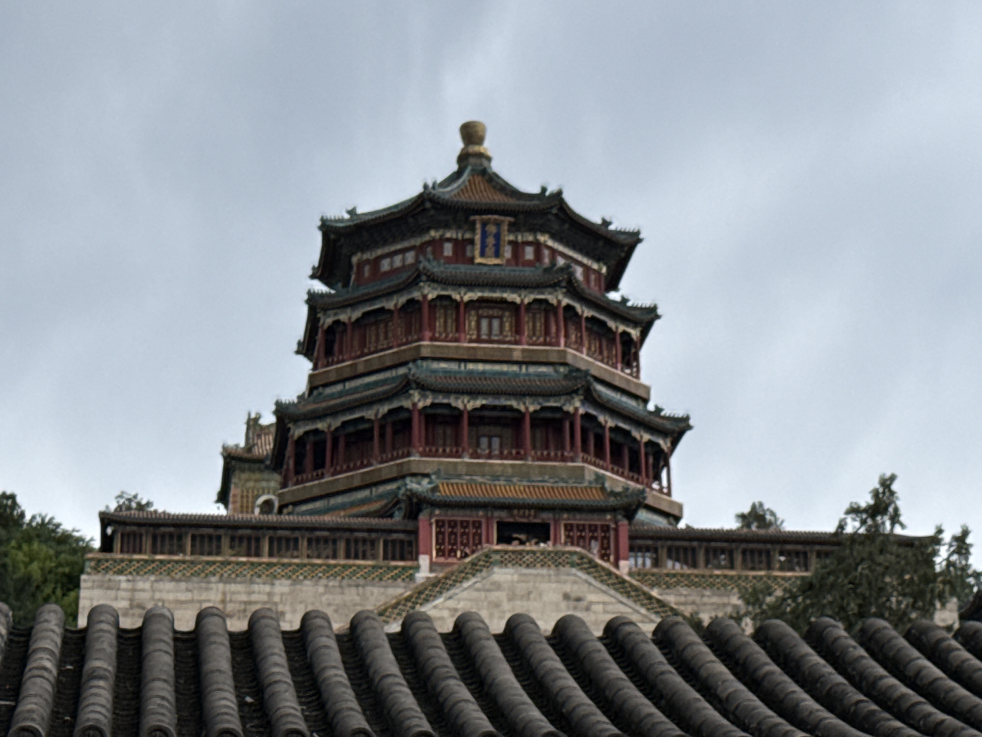 An upward-angled view captures the multi-tiered, octagonal Tower of Buddhist Incense (Fo Xiang Ge) at the Summer Palace (Yihe Yuan), situated in Haidian District, Beijing, China. The pagoda-style structure features dark red pillars, green and blue tiled roofs with distinctive upturned eaves, and intricate detailing. A golden finial adorns the apex, below which a blue plaque with golden characters is visible on the topmost visible level. Several open levels of the tower are discernible, with multiple individuals seen looking out from the balconies or windows on one of the lower levels. In the immediate foreground, the dark, curved tiled roof of another traditional Chinese building is prominent, occupying the bottom portion of the frame. Between this foreground roof and the main tower, a stone wall with a green tiled border and a wooden balustrade is visible, partially obscured by green foliage from trees on both sides. The sky is uniformly overcast and light grey, indicating a cloudy day.