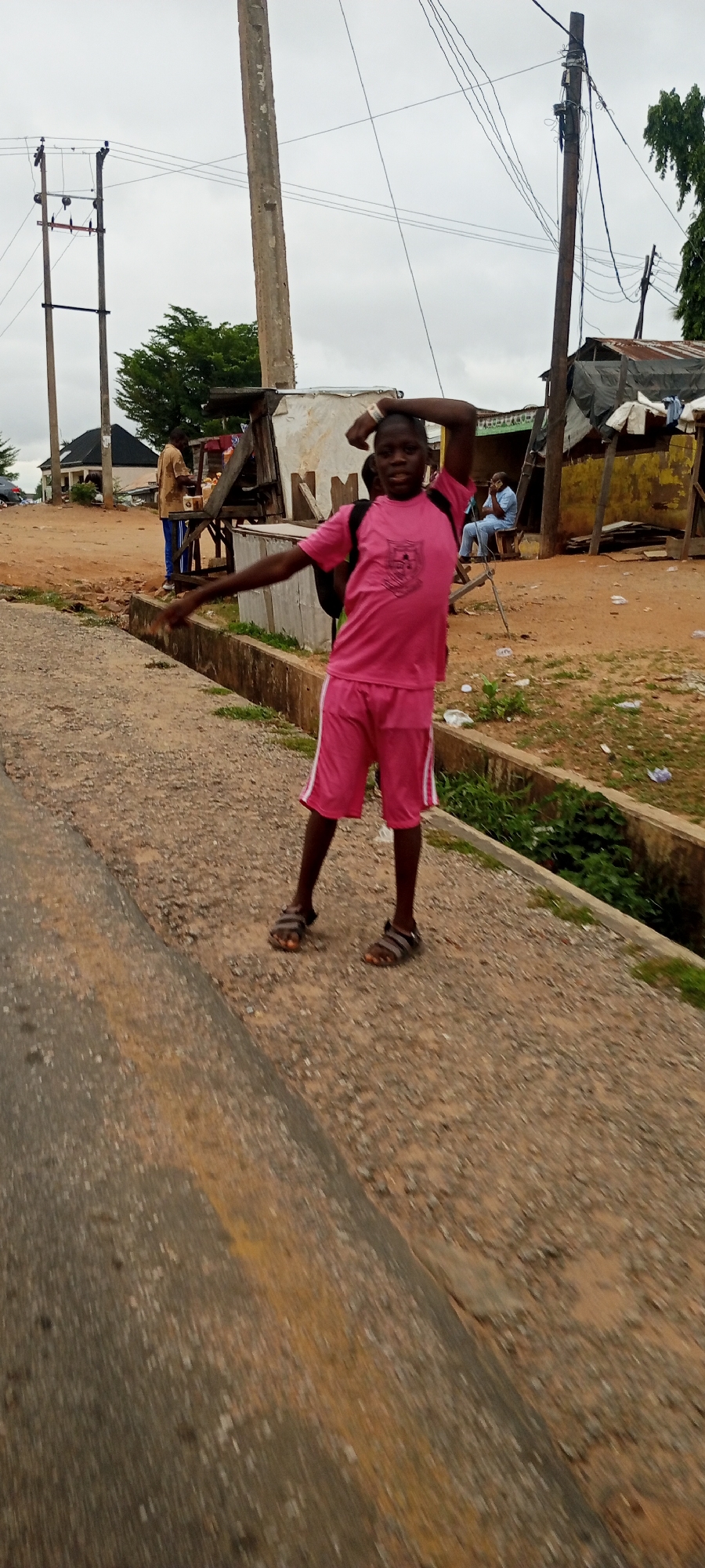The image depicts an outdoor scene, likely in Adankolo, Nigeria, with a young boy in the foreground as the primary subject. He is wearing a bright pink t-shirt and matching pink shorts with white trim, and sandals. The boy is striking a pose with his right arm raised behind his head and his left arm extended horizontally. He has a backpack on his back. Behind the boy, on the right side of the image, are several makeshift structures with corrugated metal roofs and wooden supports, characteristic of informal market stalls or housing. A man in a light blue shirt and pants is seated in one of these structures. To the left of the boy, a man in a brown shirt and blue pants is standing near a wooden stall with goods displayed. The ground is a mix of dirt and gravel, with a concrete drainage channel running along the side. Utility poles with overhead wires are visible in the background, indicating the presence of infrastructure. The sky is overcast. The overall impression is of a busy, informal urban environment.