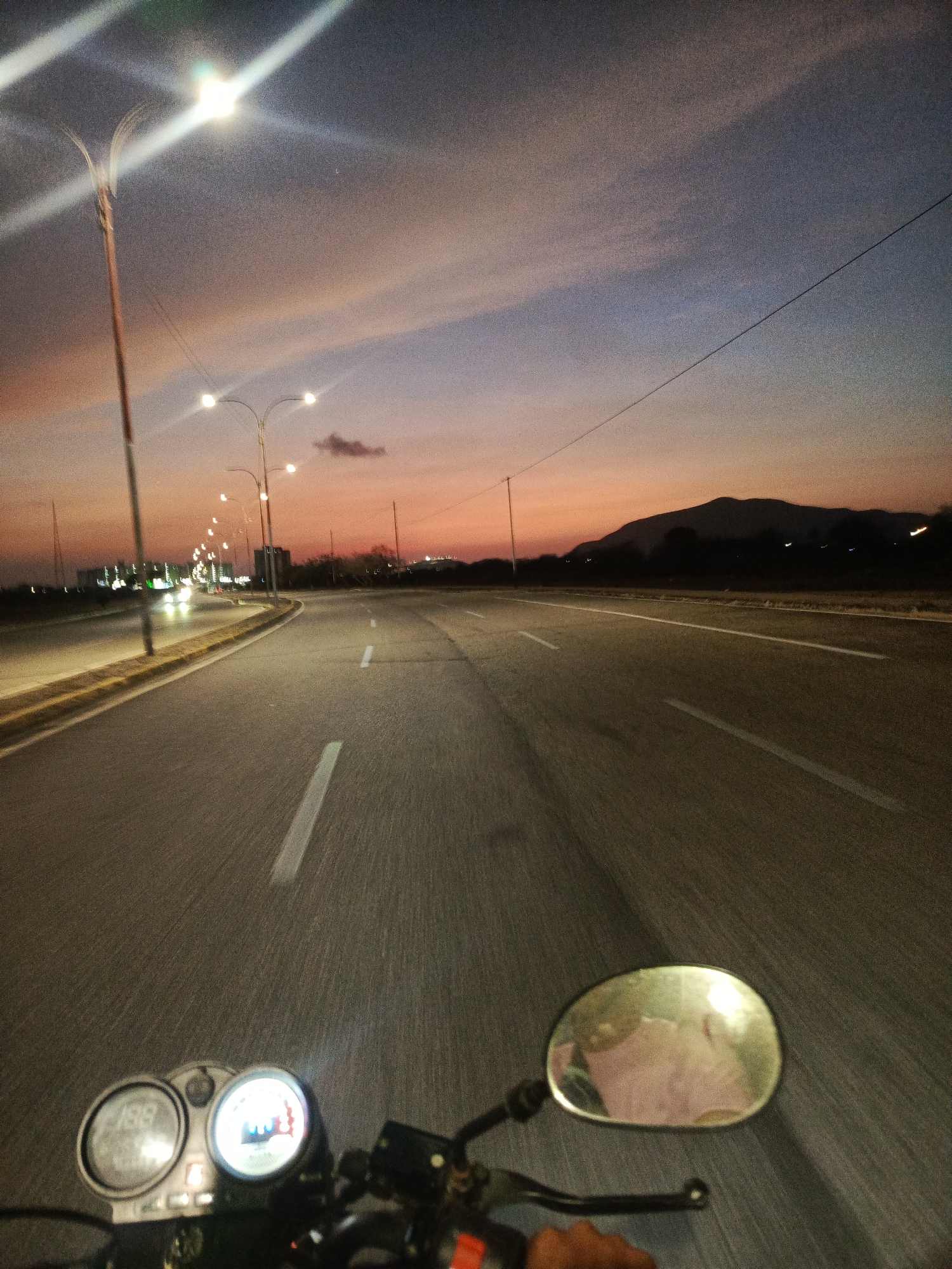 The image shows a first-person view from a motorcycle driving on an empty road at dusk in Altagracia, Venezuela. The sky is a blend of orange, pink, and purple hues. Streetlights line the road on the left, illuminating the asphalt. A mountain range is visible in the distance to the right. The motorcycle's gauges and a side mirror are in the foreground.