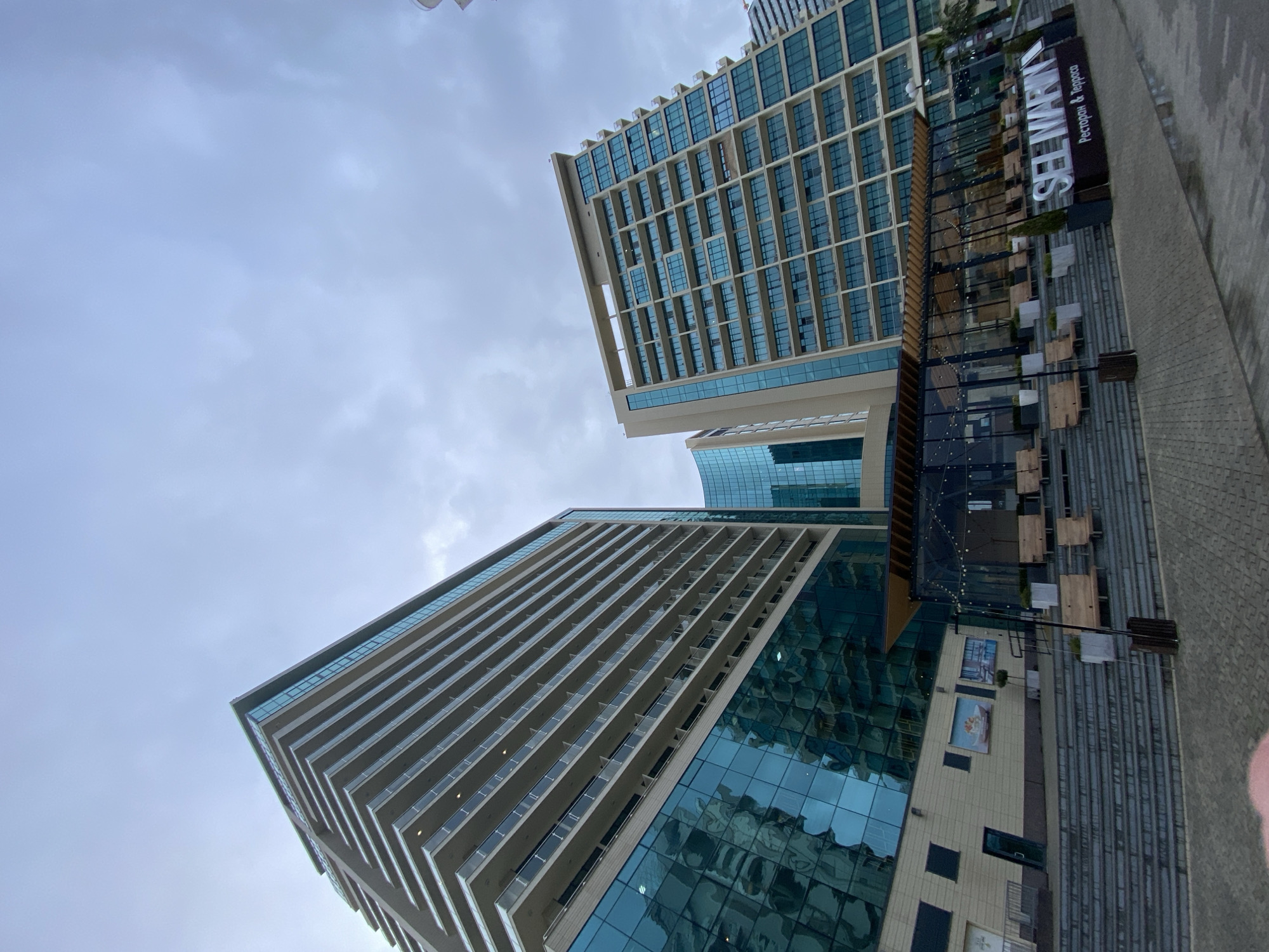 The image captures a low-angle perspective of modern architecture in Sochi, Russia. Two prominent high-rise buildings with glass facades are visible, their heights accentuated by the upward camera angle. The building on the left is distinguished by its numerous balconies. Adjacent to the buildings is a structure with a terrace and a sign reading "See Marina," presumably a restaurant or similar establishment. The sky above is overcast, contributing to the photograph's muted tones. The image is rotated to the right.