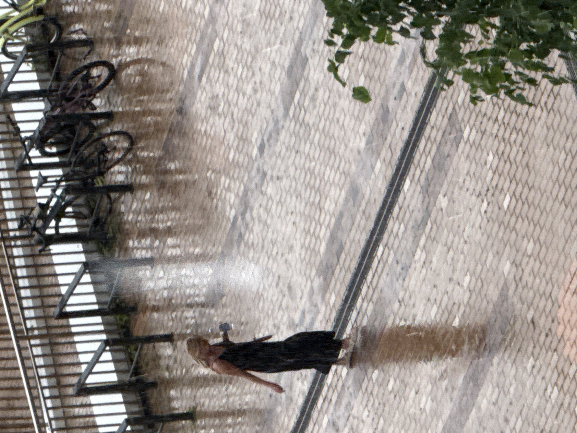 A woman in a dark, sleeveless dress and light-colored shoes walks on a wet, brick-paved street. She is holding a clear plastic cup in her right hand. It appears to be raining, as indicated by the water droplets visible in the air and the wet surfaces. In the background, there is a metal fence and several bicycles parked. The scene suggests an urban environment, possibly a public space like a park or street in Moscow, Russia, during inclement weather.