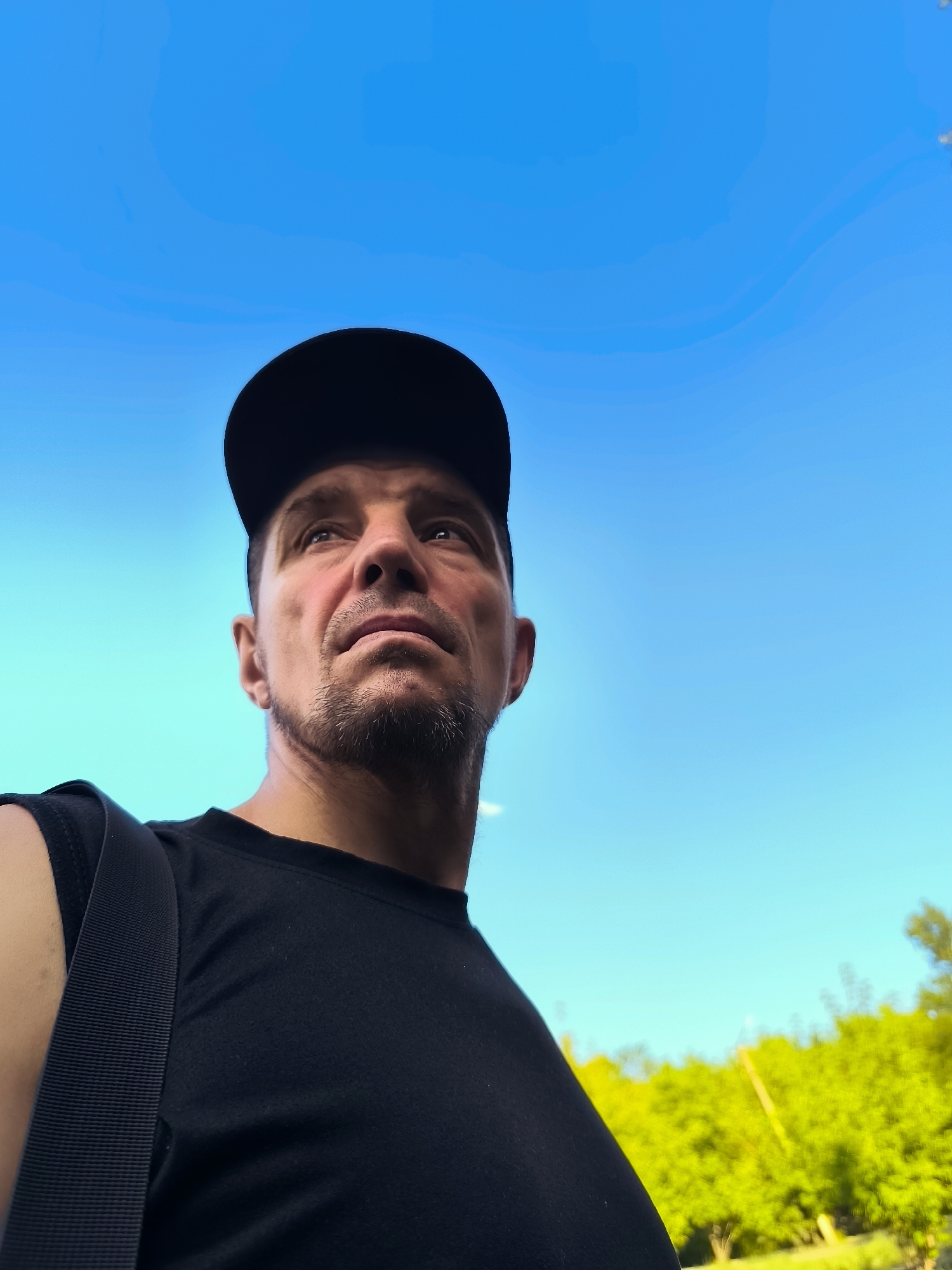 The image is a low-angle, waist-up shot of a man wearing a black t-shirt and a black baseball cap, looking upwards against a bright blue sky. He has a light beard and appears to be outdoors. A black strap, likely from a bag, is visible across his left shoulder and chest. In the background, out of focus, is lush green foliage, suggesting an outdoor environment. The image does not provide any specific visual cues to indicate the location in Qarağandı city administration, Kazakhstan.