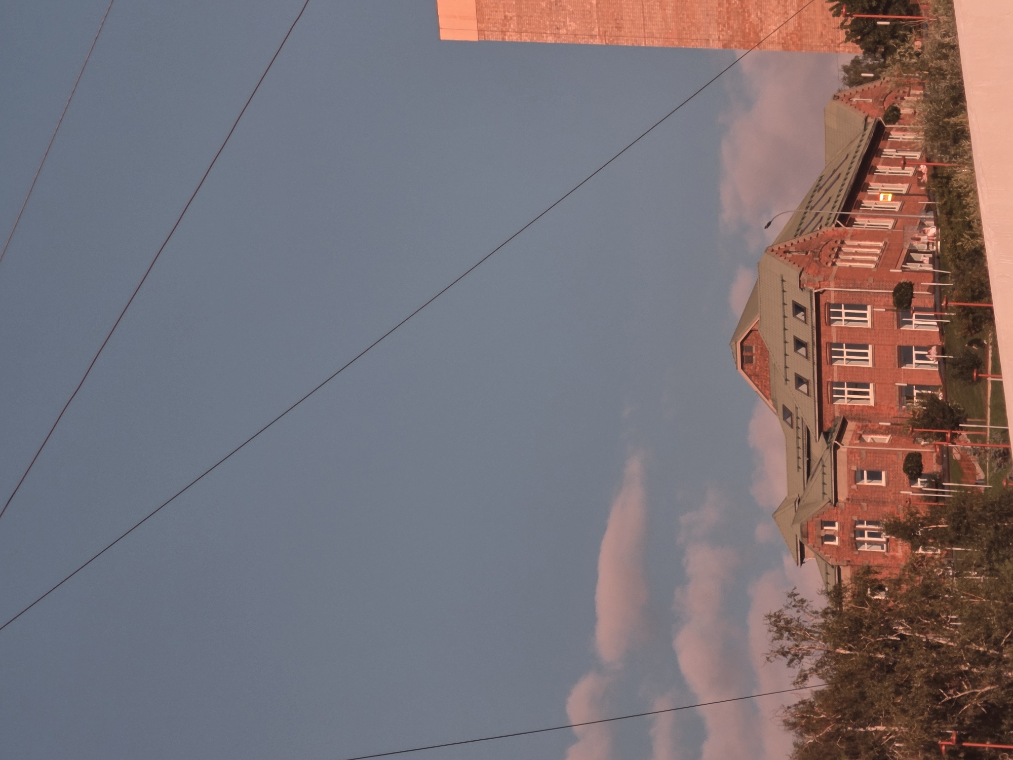The image depicts a low-angle shot of a brick building with a green roof under a clear blue sky. Several power lines traverse the upper portion of the frame. In the foreground, the tops of trees are visible. To the right, a tall brick structure extends upwards. The building appears to be a school or similar institutional building, with rows of windows and architectural details consistent with older construction. Some people are visible near the building's entrance and on the grounds. The scene suggests an outdoor setting, possibly in an urban or suburban area.