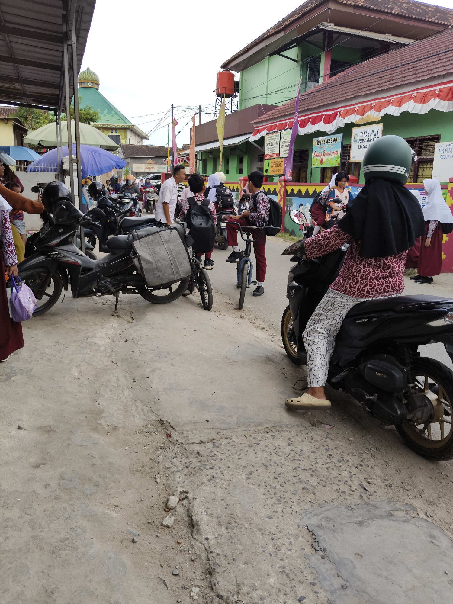 A group of people, some of whom are students in uniform, are gathered on a street in front of a school building. Several motorcycles are parked on the side of the road. In the background, a mosque with a green dome is visible. The scene appears to be in Bandar Lampung, Indonesia, as suggested by signage that includes "KOTA BANDAR LAMPUNG" and "TANAH MILIK PEMERINTAH KOTA BANDAR LAMPUNG." A woman wearing a black hijab and a patterned top is seated on a motorcycle, facing away from the camera. Other individuals are standing or walking, some carrying backpacks. The street surface is paved with concrete, showing some signs of wear and tear. Flags and banners are displayed along the school building, indicating some form of event or celebration. The weather appears to be overcast.