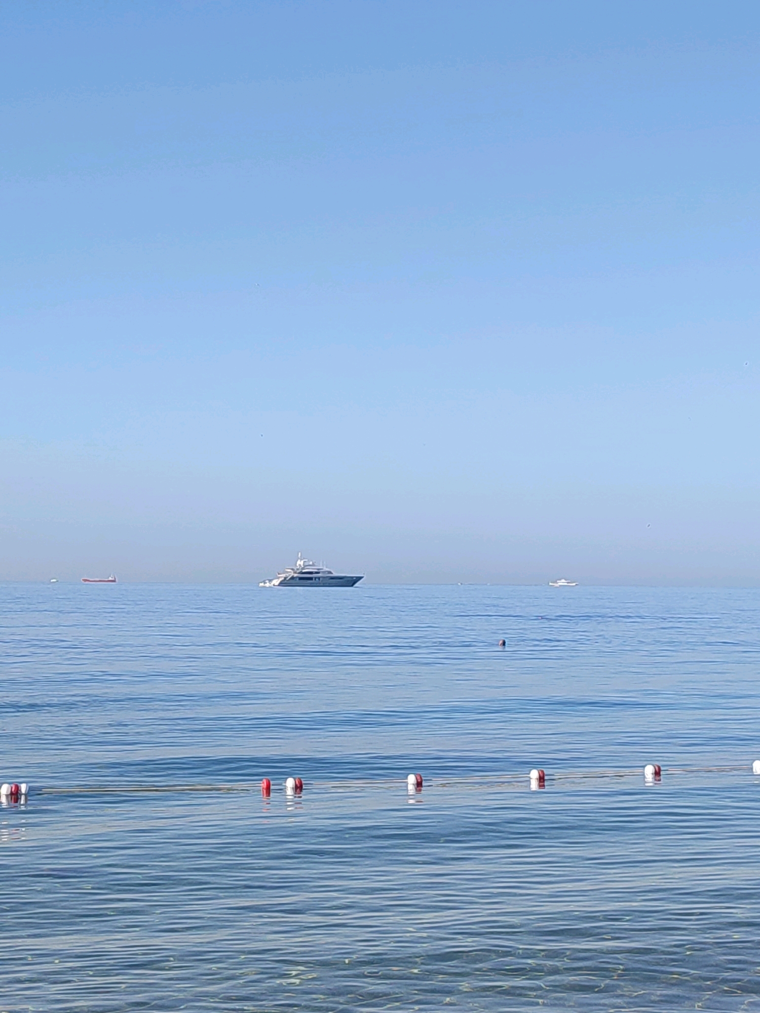 The image shows a calm, blue sea under a clear sky. In the distance, a large white yacht is visible on the water, along with a smaller red boat. A line of buoys with red and white markings floats on the surface closer to the shore. The water is clear, revealing the sandy bottom beneath. The scene suggests a serene day at the beach, possibly in Kadıköy, Turkey, given the context provided.
