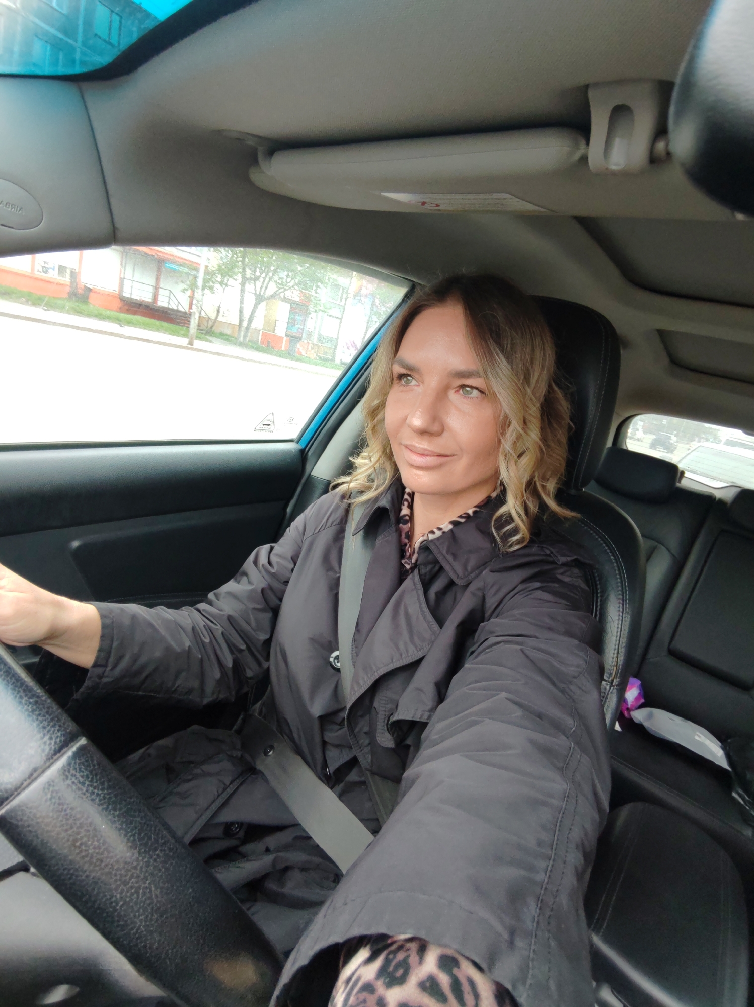 A woman is captured in a selfie-style photograph from the driver's seat of a car, set in Petropavlovsk-Kamchatsky, Russia. She has shoulder-length wavy blonde hair and light eyes, with a pleasant expression as she looks towards the left side of the frame. She is wearing a black trench coat over a leopard print top, with the seatbelt fastened securely across her chest. Her left hand rests on the steering wheel, indicating she is either driving or preparing to drive.
The car's interior features black leather seats, a sunroof, and a sun visor. Visible text includes "AIRBAG" (with "AGRIA" also partially visible, likely part of the same label) on the passenger-side A-pillar. A small vehicle icon with some unreadable text is also present on the side window.
Through the car windows, an overcast daytime street scene is visible. The external environment shows a road, buildings with distinctive reddish lower sections, and trees, suggesting an urban or suburban setting. The diffused lighting indicates cloudy weather.