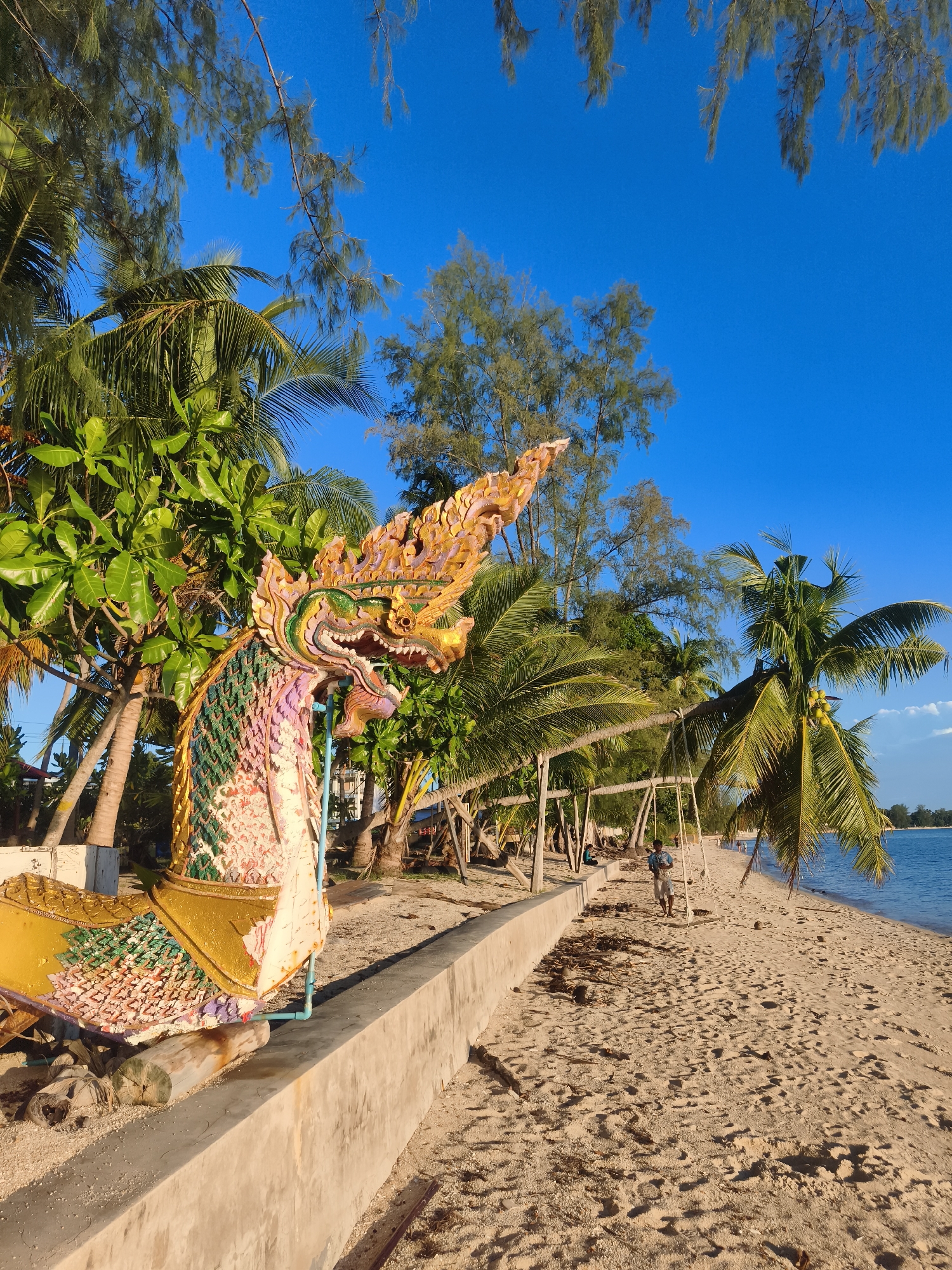 A vibrant, brightly colored statue of a mythical Naga serpent, with an ornate headdress and scales, stands on a sandy beach in Baan Lipa Noi, Thailand. The serpent is positioned in the foreground, partially obscuring a lush green tree and palm trees. Behind the statue, a concrete wall runs along the edge of the beach, separating it from a path lined with more palm trees. In the distance, several swings hang from wooden frames, and a lone person walks along the shoreline. The ocean, a calm blue, stretches to the horizon under a clear, bright blue sky. The sand on the beach is light brown and scattered with small debris. The overall scene depicts a tropical beach setting with a touch of cultural artistry.