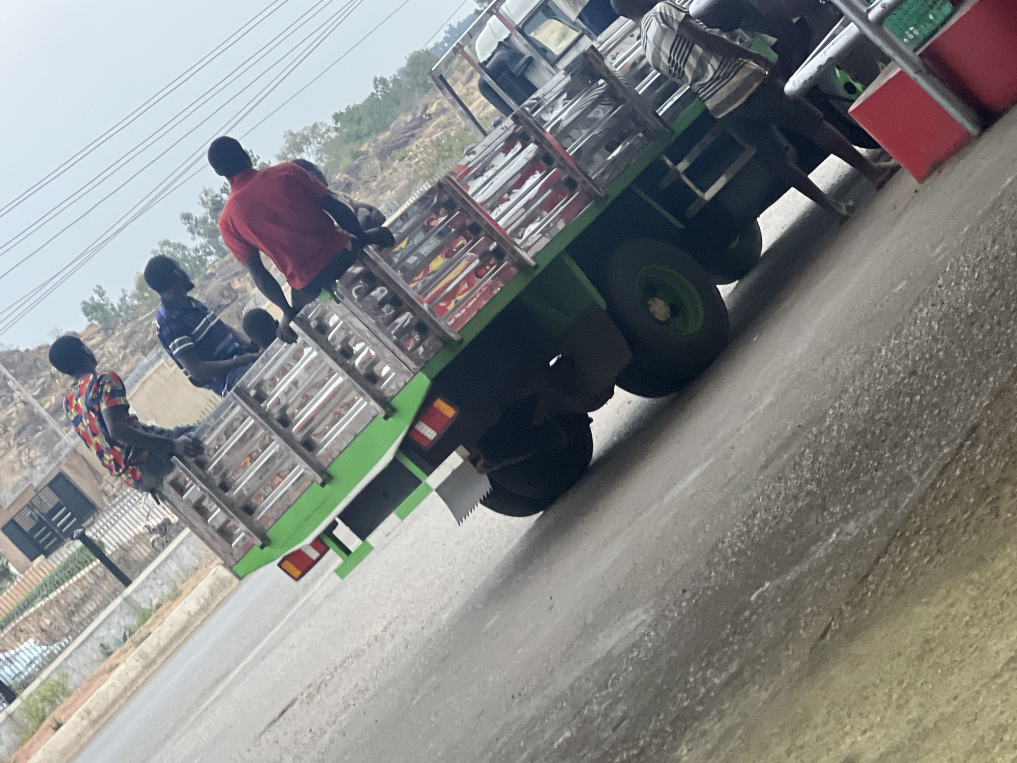 An outdoor photograph, taken from a slightly elevated and tilted perspective, captures a green and black flatbed truck transporting several men on a road in Gura Top, Nigeria. The truck features a metal cage structure over its bed, designed for carrying passengers or cargo, and has distinctive green wheels. Red and white tail lights are visible on the rear, along with a serrated mud flap.
Multiple men are riding in the back of the truck. One man, seen from behind, wears a red shirt. To his left, another man is dressed in a blue and white striped shirt. Further left, a man in a vibrant, multi-colored patterned shirt (red, blue, and yellow) is seated near the truck's edge. Another man in a plaid or striped shirt is partially visible towards the front of the truck bed, and other figures are indistinctly present.
The background features a hazy or overcast sky with overhead wires crossing it. Beyond the road, the landscape consists of hills with sparse vegetation, typical of a rural or semi-urban setting. The truck is on a paved surface, with a low curb visible on the left. The scene depicts a common mode of transport during daytime hours. No discernible text is visible on the vehicle or in the environment.