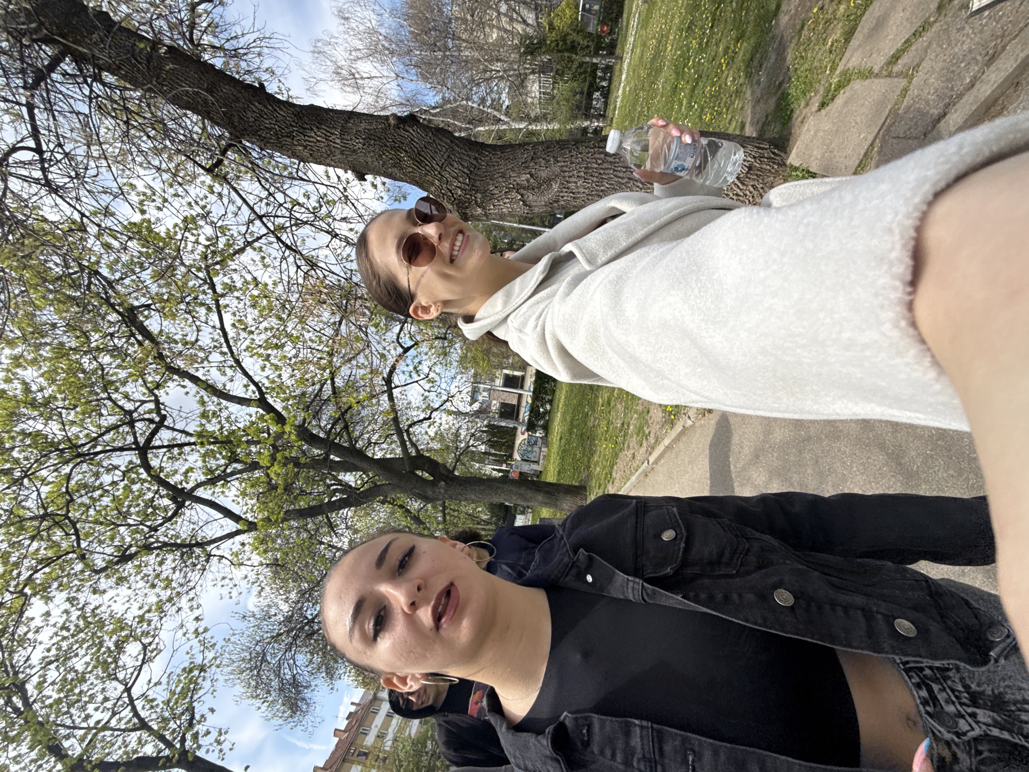 Two young women are posing for a selfie outdoors on a sunny day. The woman closer to the camera is wearing a black denim jacket and a black top, exposing part of her midriff. She has dark hair pulled back, large hoop earrings, and a chain necklace. The other woman is wearing a light-colored hoodie and sunglasses. She is holding a water bottle in her right hand. Behind them, there are trees with fresh green leaves, a grassy area with some yellow flowers, and buildings in the background. The photo appears to have been taken in a park or green space in Sofia, Bulgaria, judging by the architectural style of the buildings.