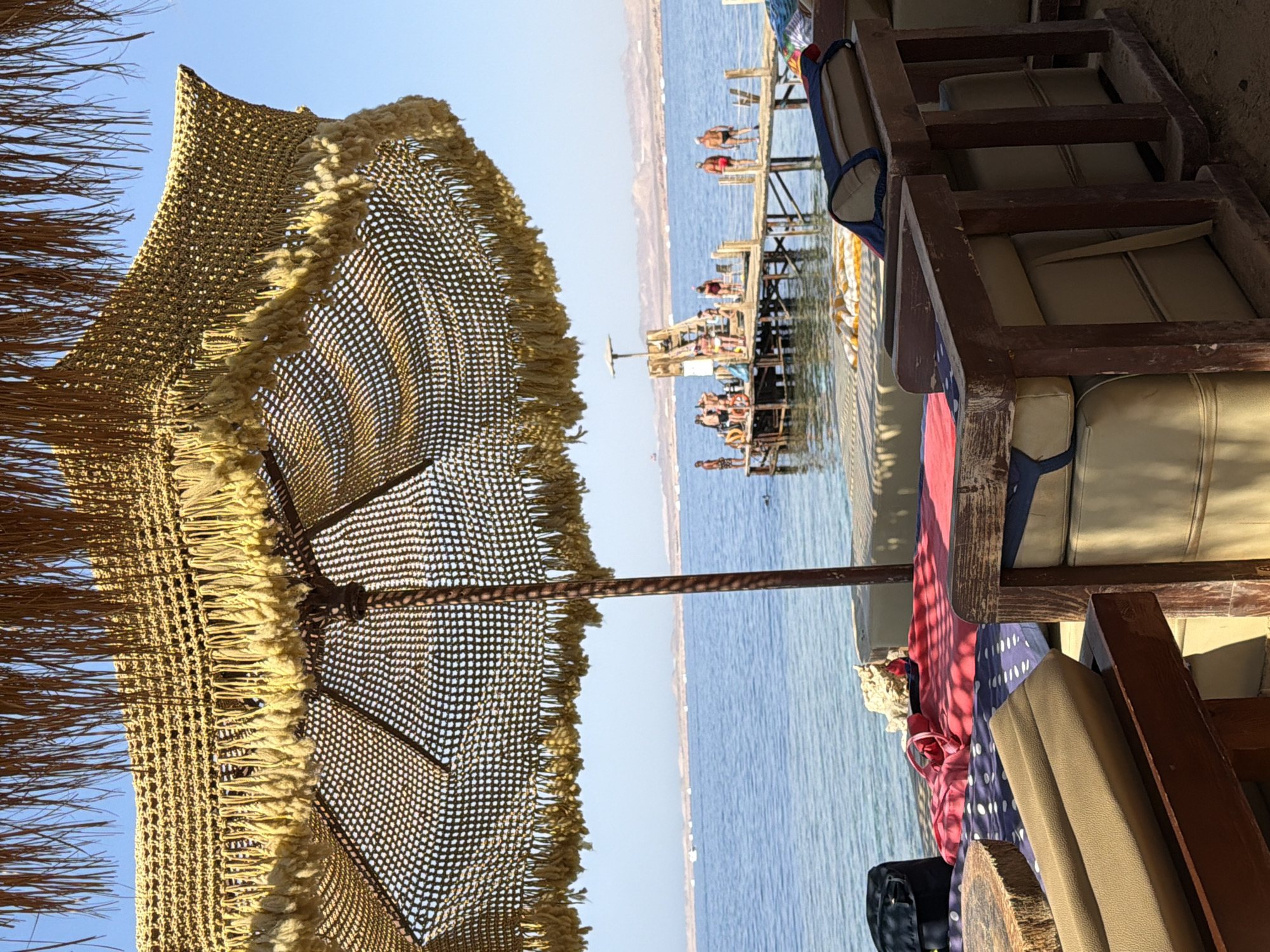 The image depicts a beachfront seating area with a thatched umbrella in the foreground, offering shade. Beyond the seating, a wooden pier extends into the clear blue sea. Several people are visible on the pier, some standing and others gathered together, suggesting recreational activity. The background shows a distant shoreline with mountains. The scene appears to be a resort or beach establishment in Sharm El Sheikh, Egypt, given the architectural style of the umbrella and the general ambiance of a coastal tourist destination.