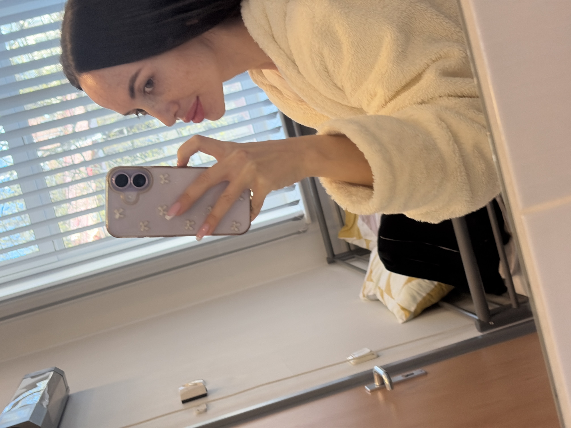 A woman with dark hair, light skin, and freckles is taking a selfie in a mirror. She is wearing a white, fluffy robe. She is holding a purple phone with a floral design. The background shows a window with white blinds. Outside the window, there are trees. The reflection in the mirror reveals a bathroom environment with a countertop, a door handle, and a pillow. There is no definitive information to confirm the location in Saint Petersburg, Russia.