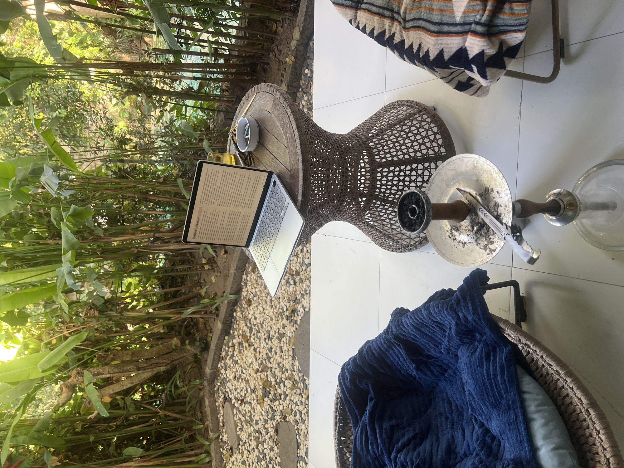 The image shows a laptop on a wooden table in an outdoor setting. The table is next to a stone garden. A hookah sits on the ground next to a brown woven chair with a blue blanket. The scene is in Kerobokan, Indonesia.