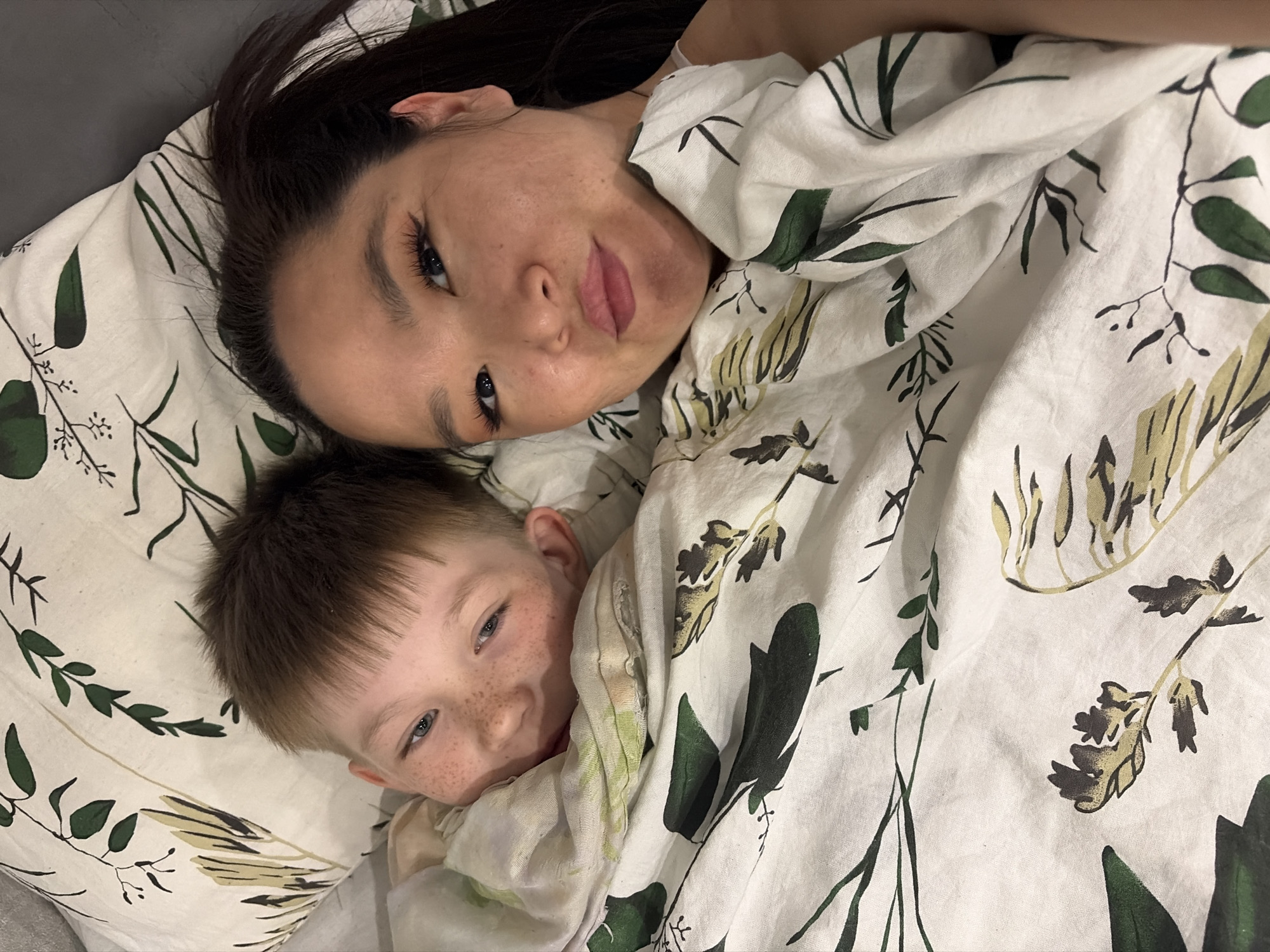 A top-down shot shows a woman and a young boy lying on a bed covered with a white floral-patterned duvet. The woman, with long dark hair, is positioned above the boy. Her face is tilted towards the camera. The boy, with light brown hair and freckles, is partially covered by the duvet, with only his face visible. Both are looking at the camera. The location is Unknown, Unknown.