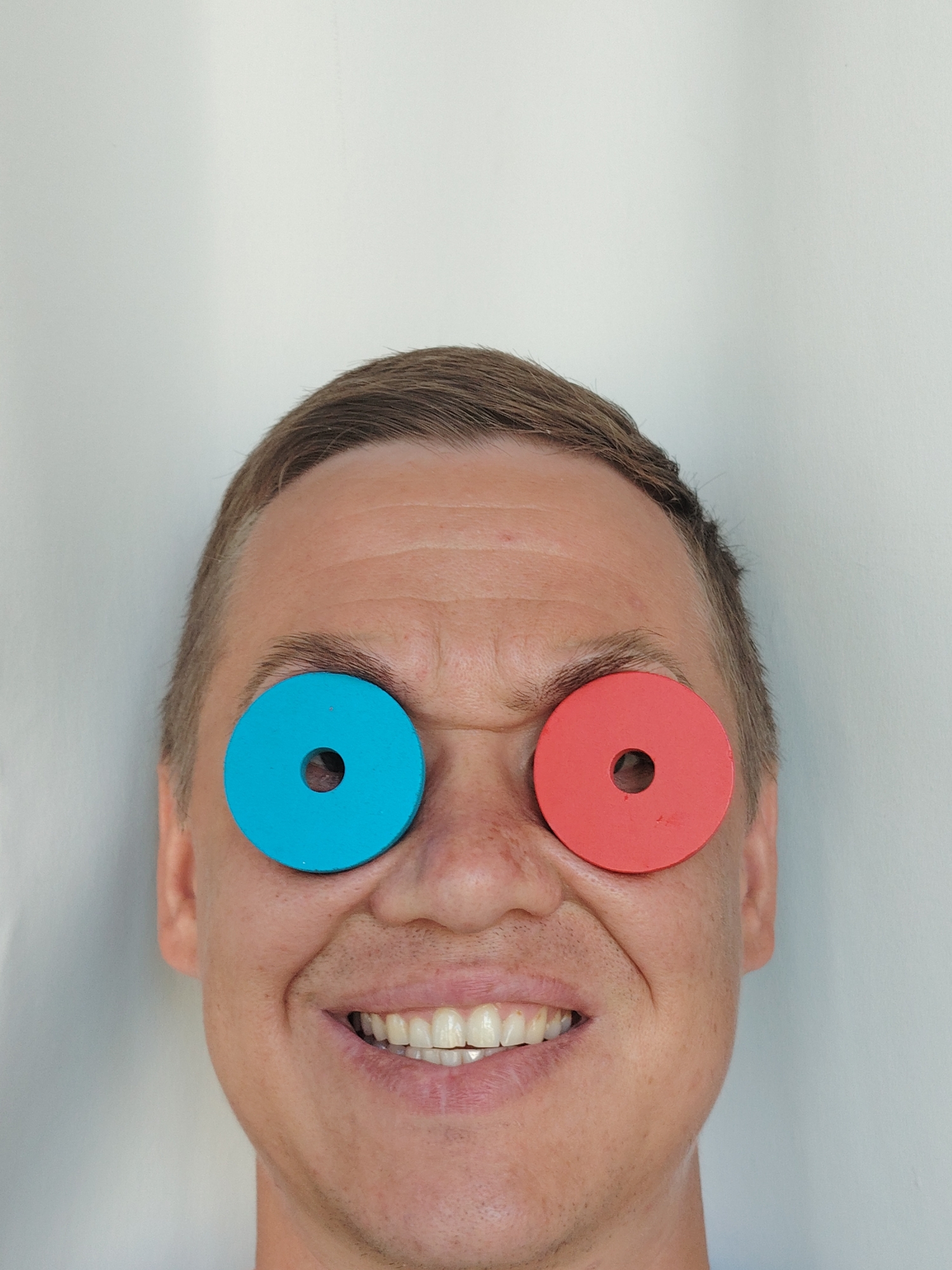 A close-up, head-and-shoulders shot captures a person's face against a plain white background. The individual, with fair skin and short brown hair, is smiling broadly, revealing their teeth. Their eyebrows are dark and arched, and there are visible wrinkles on their forehead. The most striking feature is the placement of two colorful circles over each eye, resembling large, unadorned earrings or decorative eye patches. The circle on the left is a bright cyan, and the one on the right is a vibrant red. Each circle has a smaller, perfectly round hole in its center, through which a glimpse of the person's eye can be seen. The lighting is even, casting soft shadows that highlight the contours of the face. The image is cropped tightly, focusing exclusively on the face and the unusual eye coverings. There is no visible context or environmental detail to suggest a specific location within Chaikovsky, Russia, or any other city.