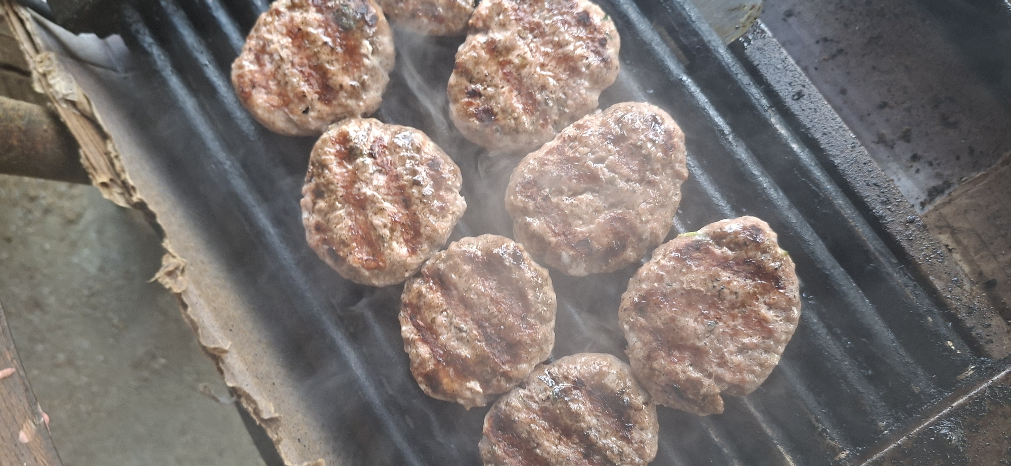 This image captures a close-up, top-down view of several grilled patties, likely made of ground meat, arranged on a black metal grill. The patties are browned with distinct grill marks, indicating they are being cooked. Wisps of steam rise from the grill, suggesting the patties are hot. The grill itself appears to be outdoors, with some weathered wooden elements visible at the top and a concrete or dirt surface below. The overall impression is of a casual outdoor cooking environment. The image does not provide any specific visual cues to pinpoint its location within Lyaskovets, Bulgaria.