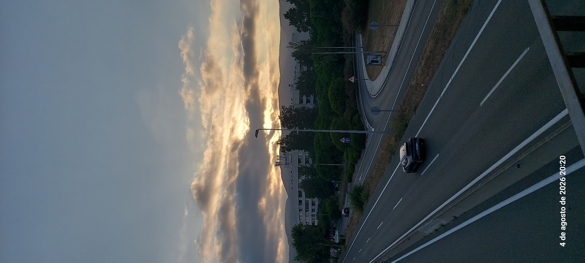 The image captures a highway scene in Castelldefels, Spain, at dusk. A dark-colored four-door sedan is visible driving on the highway, heading away from the camera. The highway is flanked by green trees and appears to be a multi-lane road with a concrete divider. In the background, several multi-story buildings are visible, along with hills beyond them. The sky is partly cloudy, with the sun setting and casting a warm glow through the clouds. A lamppost is also present on the side of the highway. The timestamp "4 de agosto de 2026 20:20" is visible in the bottom left corner of the image, indicating the date and time it was taken.