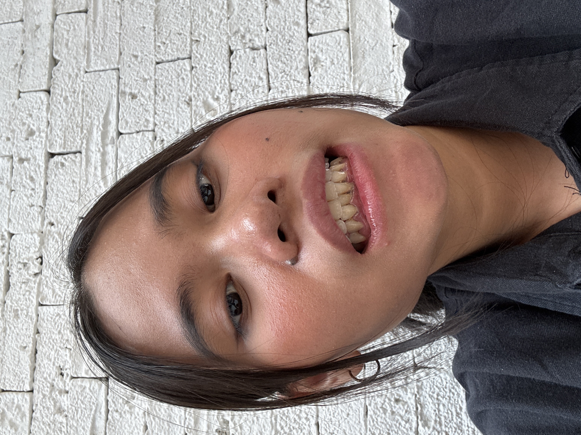 A close-up, eye-level, outdoor selfie shows a young person in South Jakarta, Indonesia, against a white brick wall. The subject, with dark hair, is wearing a black collared shirt and has makeup on, including blush and eyeliner. Their mouth is slightly open, revealing teeth that appear discolored. The lighting suggests it is daytime.
