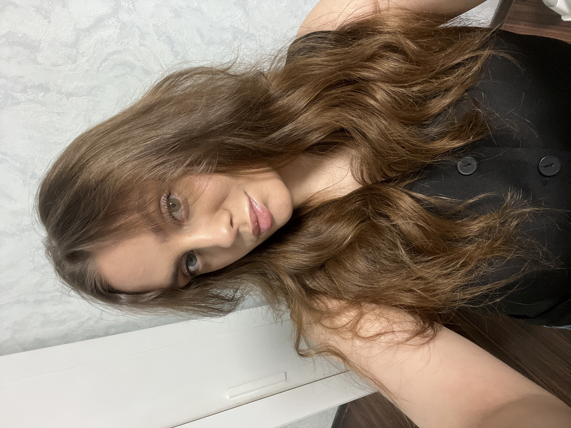 A woman with long, wavy, light brown hair is looking at the camera. She has light skin and appears to be wearing minimal makeup, with a glossy lip. She is wearing a black top with two buttons visible. The background consists of a textured white wall and a wooden floor. The image appears to have been taken indoors. There's no specific evidence within the image to indicate that the location is in the city of Chaikovsky, Russia.