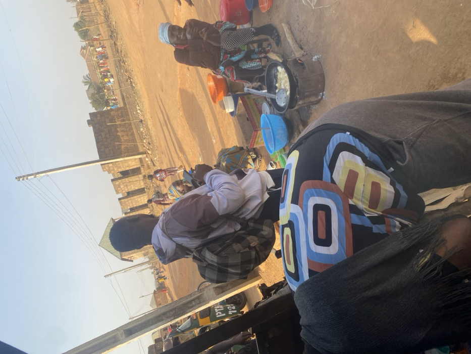 The image depicts an outdoor scene in Udu, Nigeria, characterized by a bustling roadside environment under clear daylight. In the immediate foreground, the back of an individual wearing a dark shirt with large, colorful geometric patterns and grey trousers is visible, partially obscuring the lower left of the frame.
The midground features a man standing with his back mostly to the camera, wearing a white and brown hooded sweatshirt and a plaid backpack. He holds a smartphone in his right hand. To his right, an older woman is seated on a low stool, wearing a blue headwrap, a brown top, and a brightly patterned skirt. She is smiling and looking towards the viewer's general direction. Next to her, a cooking station is active: a dark pot containing items (possibly plantains or yams) frying in oil sits on a makeshift stove, likely fueled by wood or charcoal, with visible flames. A metal slotted spoon rests in the pot. Nearby are colorful plastic bowls, including an orange and a light blue one, along with a red lidded container.
The ground across the scene is unpaved, dry reddish-brown dirt. In the background, a dirt road stretches into the distance, with several individuals, including children, walking on it. Partially constructed buildings made of concrete blocks, some with exposed rebar, line the right side of the road, separated by a fence. Utility poles with overhead power lines are visible throughout the scene. On the far left, part of a green and yellow tricycle (Keke Napep) with "PTA 721" text is discernible. The sky is bright and clear, casting distinct shadows.