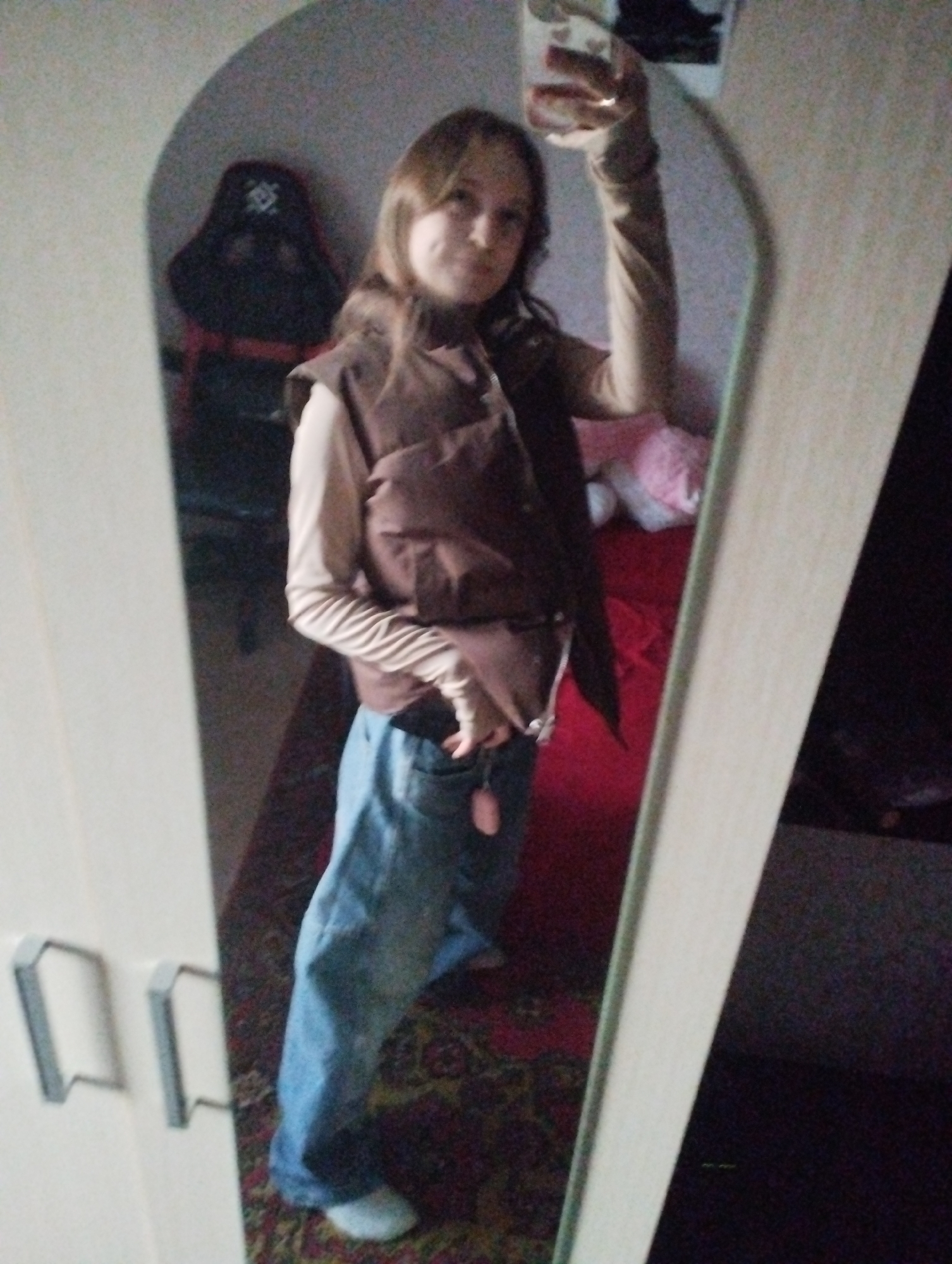 A person stands in front of a mirror, taking a selfie with a mobile phone. The person is wearing a long-sleeved tan shirt, a brown sleeveless puffer vest, and loose-fitting blue jeans, along with white socks and light-colored shoes. The background includes a black gaming chair on a red frame, a red bedspread with a pink stuffed animal, and a floral rug. The mirror is attached to a white wardrobe with silver handles. The picture was taken indoors. There is no verifiable evidence suggesting that the location is in the city of Saratov, Russia.