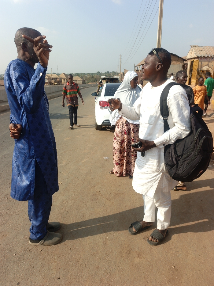 Two men stand on a street engaged in conversation. The man on the left wears a blue patterned outfit and grey shoes. The man on the right wears a white outfit, sandals, and has a black backpack. A white car is parked nearby, with a woman in a hijab standing beside it. Several other people are walking in the background. The setting appears to be a sunny day in a residential or commercial area.