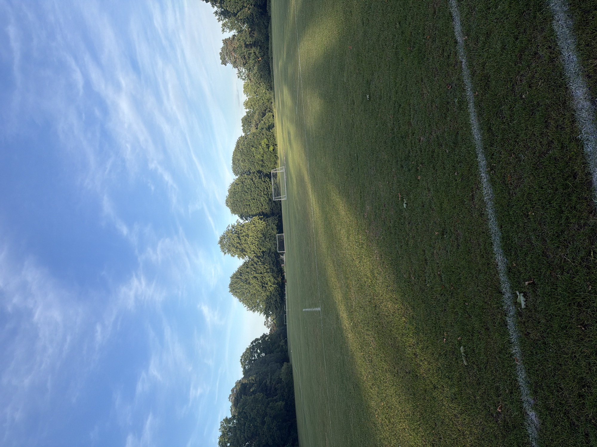 This image displays an outdoor grass sports field, likely a football pitch, situated in Maidstone, United Kingdom. The field is marked with white lines, including a prominent line across the foreground and another perpendicular line further back. Three goal frames are visible: a full-sized white goal with a net positioned on the right mid-ground, and two smaller, white or light-colored goal frames without nets, located further left and center-left on the field. The periphery of the field is lined with mature deciduous trees, exhibiting dense green foliage. Above, the sky is clear blue with sparse, wispy white clouds. Long shadows from an unseen source on the left extend across the grass, indicating a low sun angle. No individuals, animals, or active events are present within the frame. Scattered dry leaves are visible on the grass in the foreground.