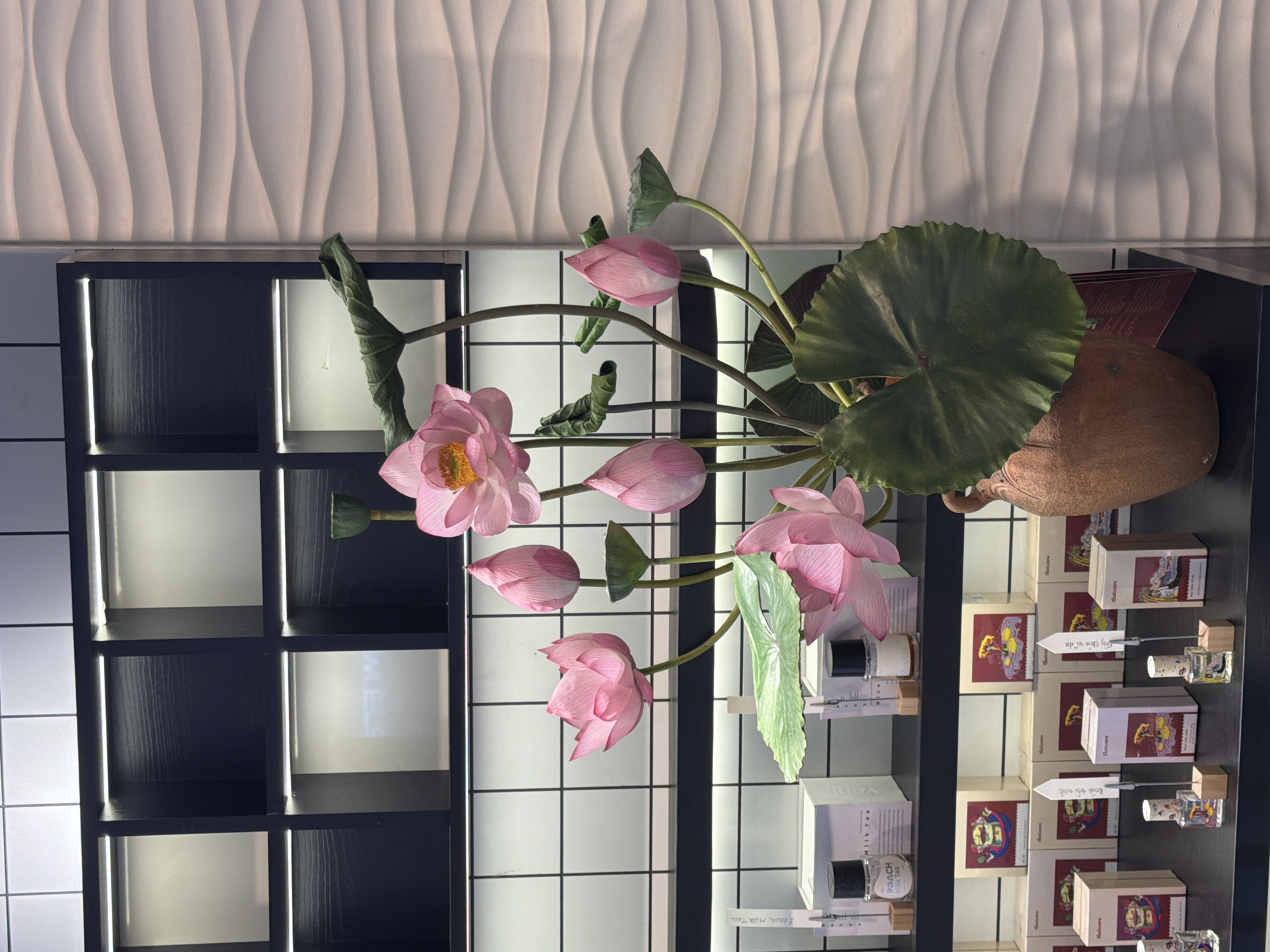 The image depicts a still life arrangement featuring a bouquet of pink lotus flowers and leaves in a terracotta vase. The background consists of a wall with a textured, wavy pattern to the right and a black grid shelving unit with illuminated compartments to the left. The shelves below the grid are stocked with various products, including small bottles and boxes, suggesting a retail or display setting. The overall lighting is soft and diffused. The scene is likely located in a shop or showroom in Nha Trang, Vietnam, a coastal city known for its lotus flowers, which are culturally significant in Vietnamese tradition.