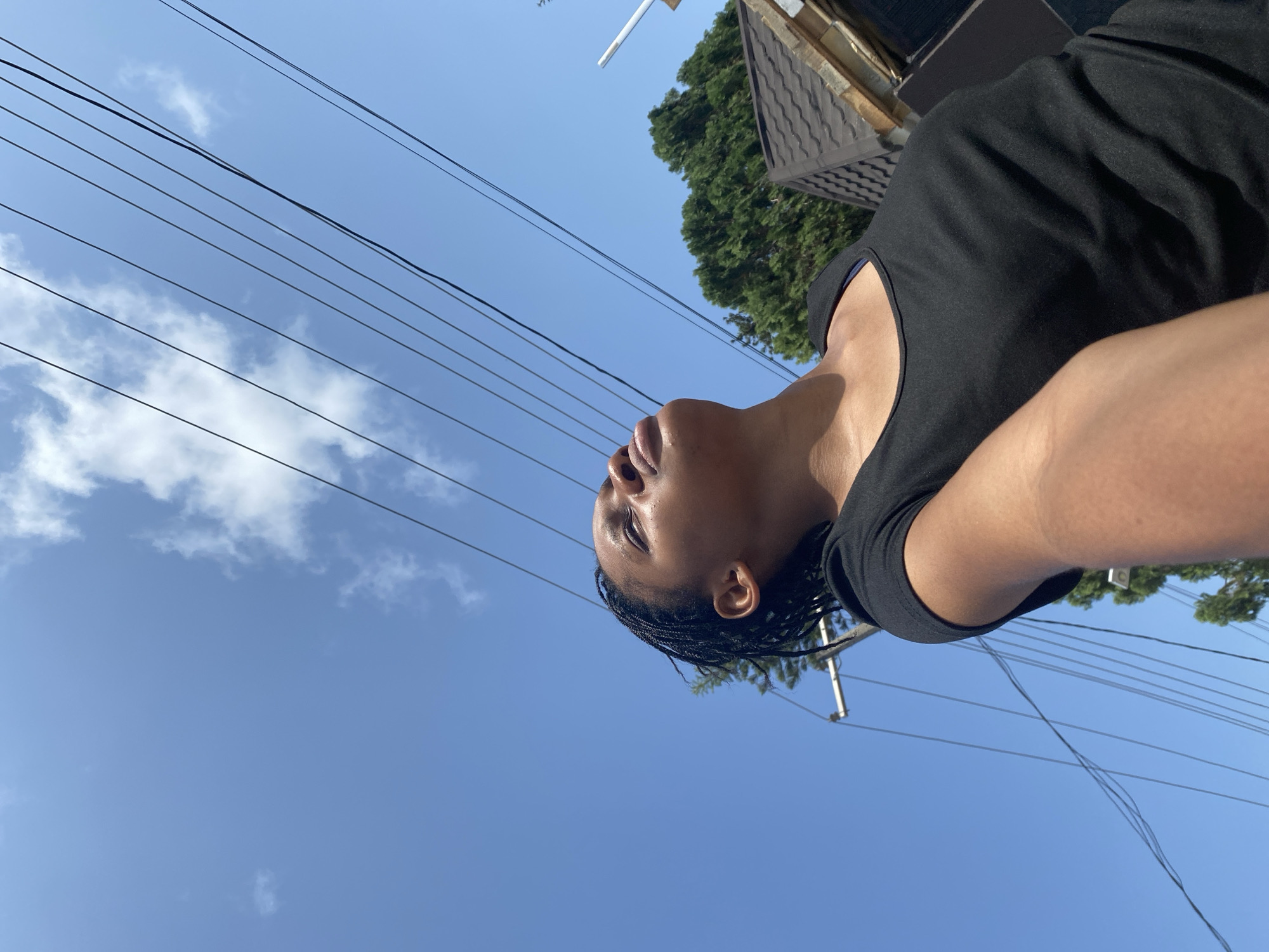 A low-angle, daytime shot shows a person with brown skin and braided hair, wearing a black shirt. The person is looking upward. The background features a blue sky with light clouds, power lines, a green tree, and part of a building with a roof. The location is Iranla, Nigeria.