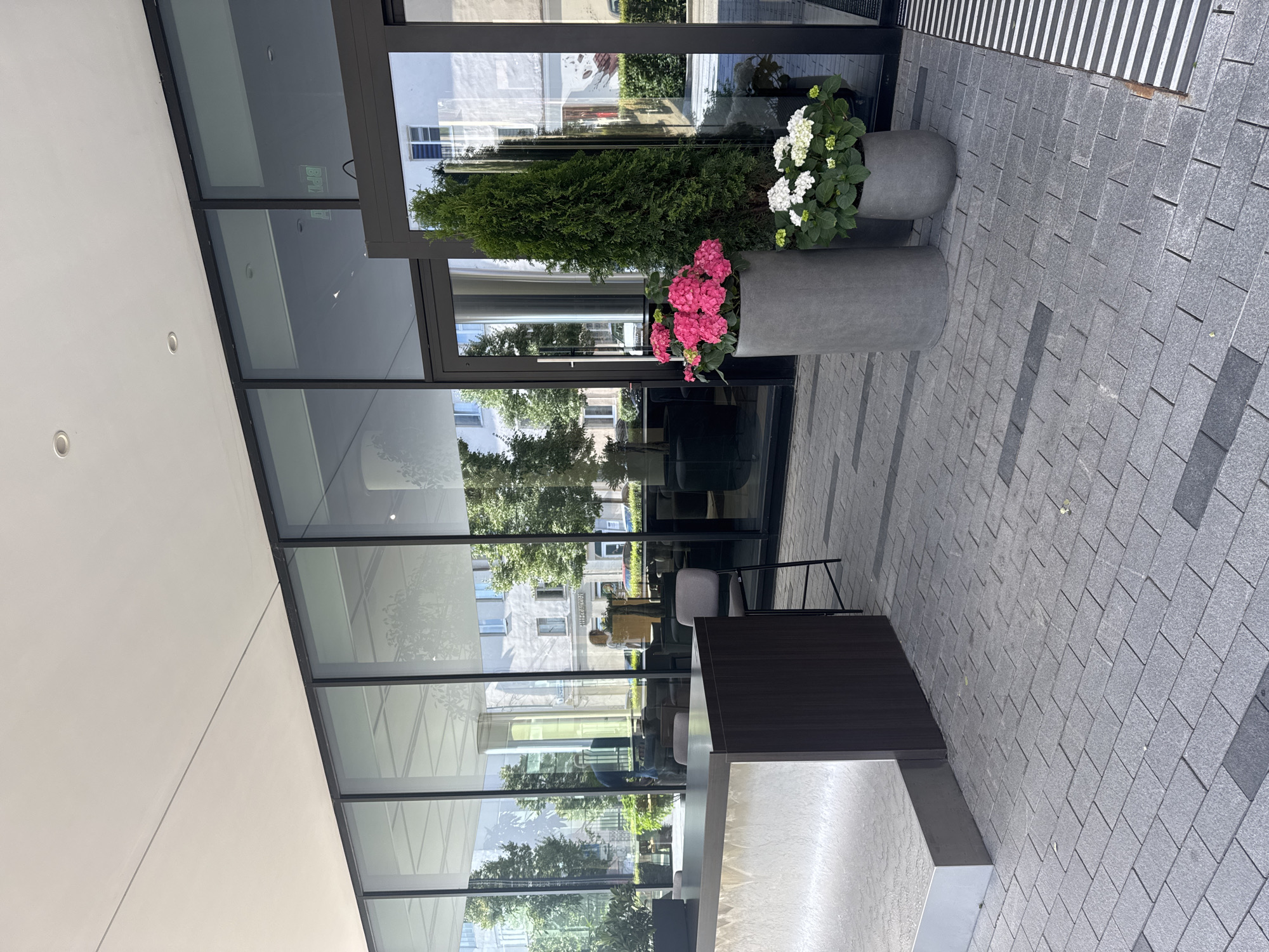 This image shows an outdoor seating area in front of a modern building. The scene is viewed from a slightly elevated perspective, with the camera angled downwards and to the right. Two large, gray planters with decorative plants are prominently featured. One planter contains a tall, green evergreen tree, and the other holds blooming pink and white hydrangeas. The building has large glass windows that reflect the surrounding environment, including trees and other buildings.
In the reflection on the glass, an interior space can be seen, furnished with tables and chairs, suggesting a cafe or restaurant. A dark wood desk or counter is in the foreground, possibly an information or reception desk. The ground is paved with gray rectangular tiles arranged in a patterned walkway. The lighting suggests it is daytime, with bright sunlight casting shadows. The overall setting appears to be a clean, well-maintained urban environment, likely a commercial or public space. There is no visible text in the image.