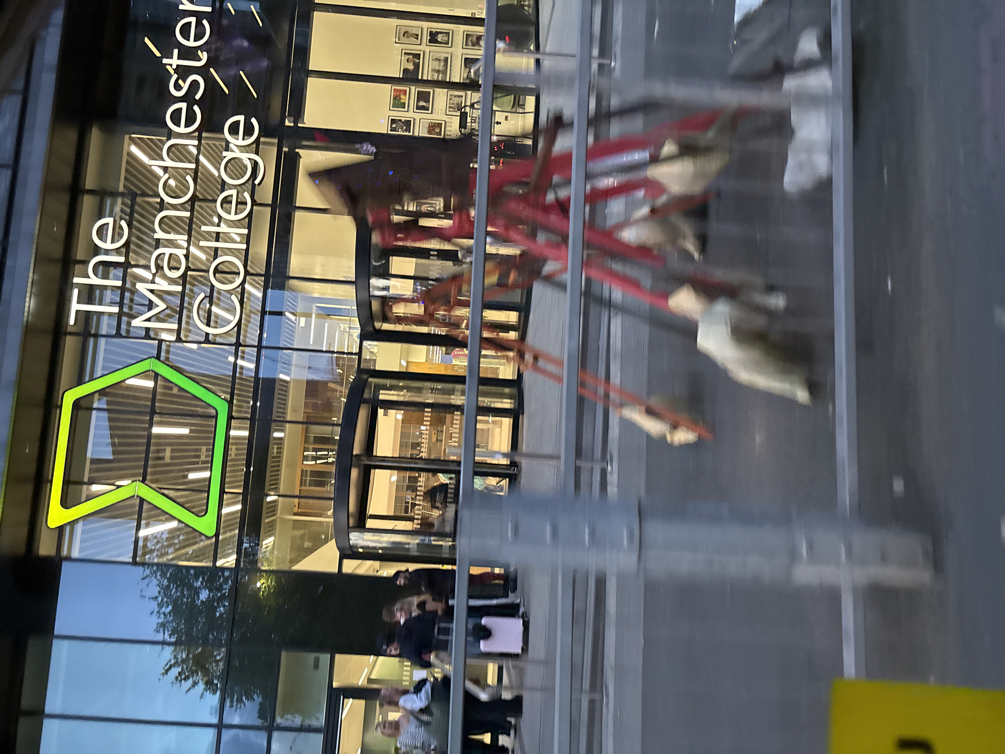 The image captures the exterior of The Manchester College in Manchester, United Kingdom. The college's signage is prominently displayed, featuring the name "The Manchester College" in white lettering with a green logo. The building's façade is predominantly glass, reflecting the surrounding environment. A group of people is visible near the entrance, some with luggage, indicating potential students or visitors. A metallic barrier is in the foreground.