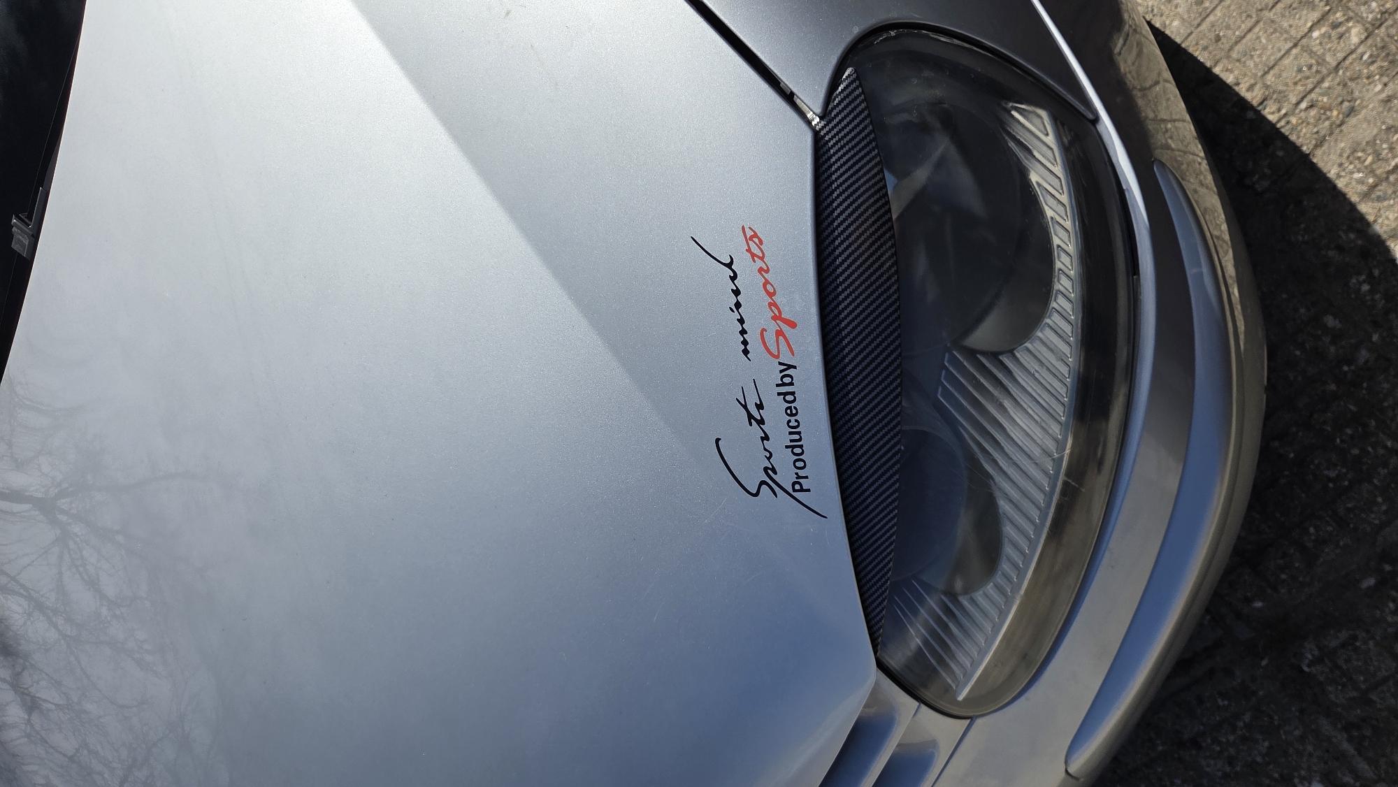 A close-up, slightly angled, outdoor daytime shot of the hood and headlight area of a silver car. A decal featuring the words "Sports mind Produced by Sports" in stylized black and red font is visible on the hood. A strip of carbon fiber-look material is placed next to the headlight. The headlight itself is large and curved. The background shows a blurred concrete surface. The image's location is not discernible as Bania, Bulgaria.