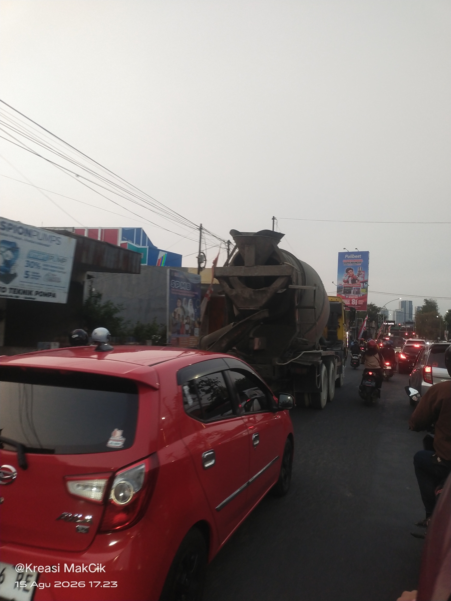 The image depicts a street scene in Surabaya, Indonesia, likely during daylight hours given the illumination. The foreground is dominated by a red car, possibly a hatchback, with a visible license plate starting with "6 PE". On the roof of this car, two helmets are placed. Behind the red car, traffic is heavy. A large cement mixer truck is prominent in the middle ground, with its drum clearly visible. Several motorcycles and cars are present in the traffic, with some riders visible. In the background, buildings and billboards are visible, including one for "Polibest" and another advertising "SPIONPUMPS". The sky is overcast. The scene appears to be on a road with multiple lanes and utility poles with overhead wires. The timestamp on the image indicates it was taken on August 15, 2026, at 17:23.
