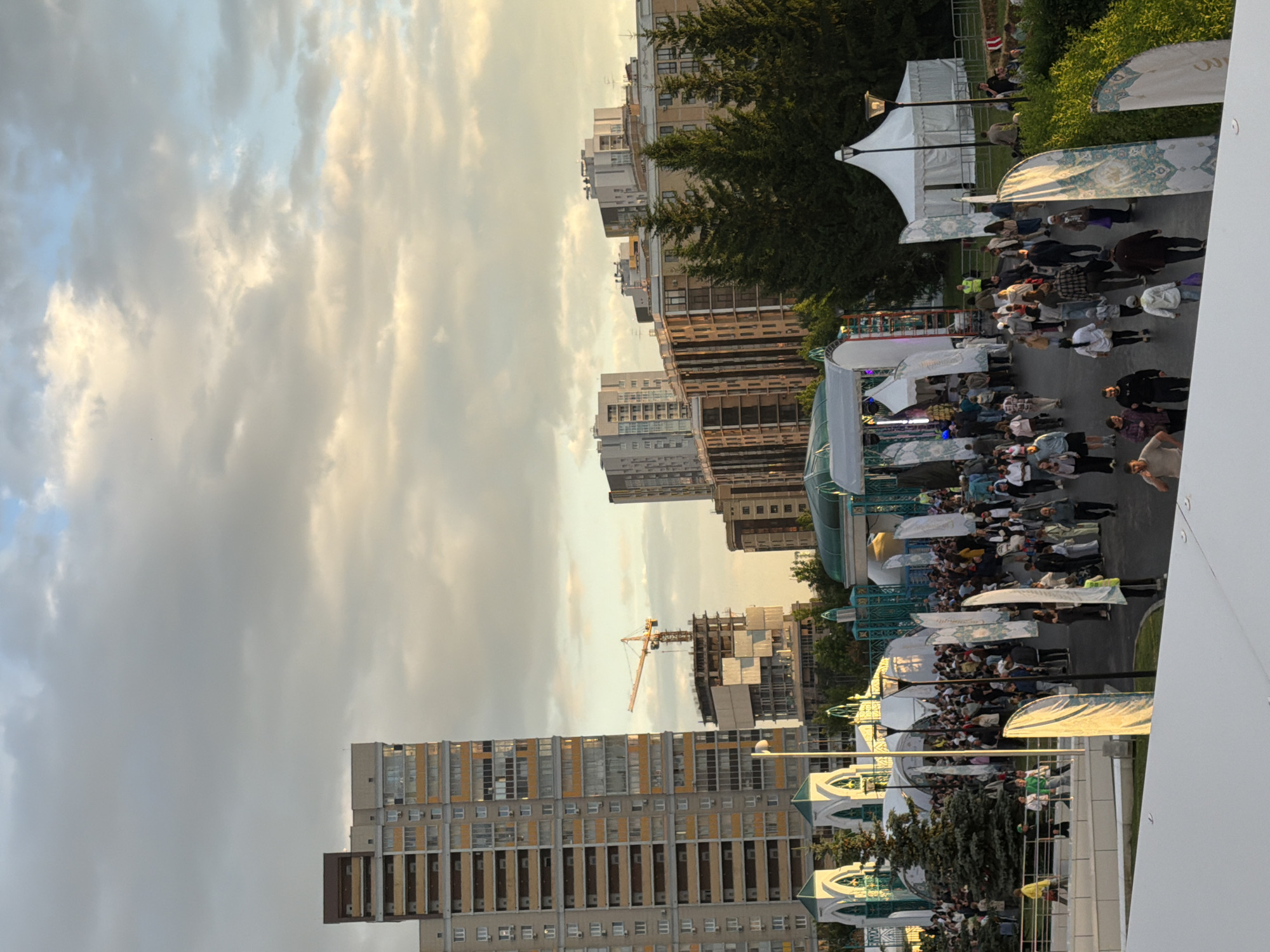 This image depicts an outdoor event with a crowd of people gathered. In the background, several tall apartment buildings and construction sites are visible, suggesting an urban environment. The foreground is occupied by a walkway with tents, banners, and people milling about. The sky is partly cloudy, with the sun casting warm light, indicating it might be late afternoon or early evening. There are trees and green spaces interspersed with the man-made structures, suggesting a park or an open area within a city. The people are dressed in casual attire, and some appear to be walking, talking, or observing the surroundings. The overall scene conveys a sense of community gathering and activity.