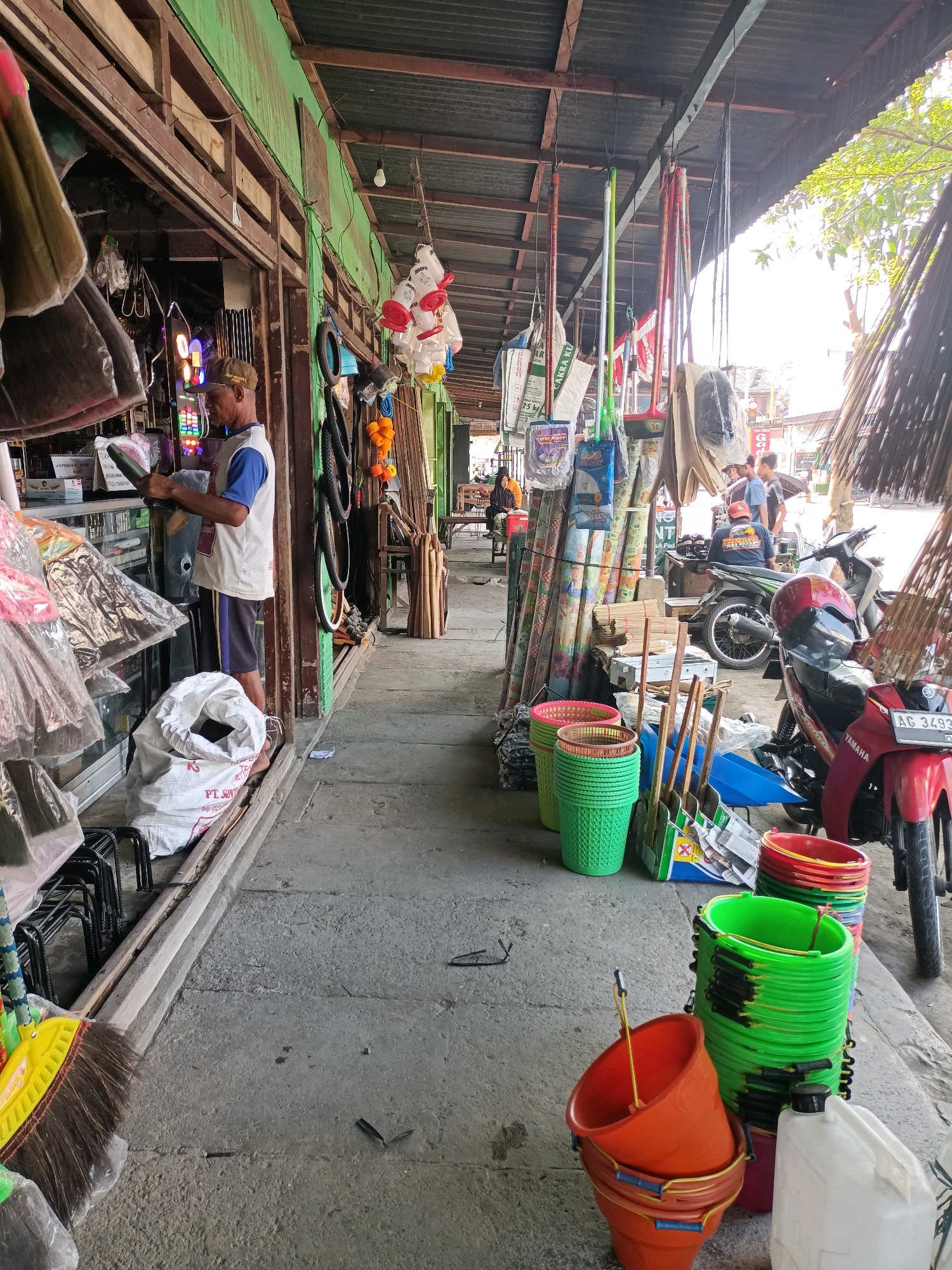 The image depicts an outdoor market stall in Badas, Indonesia, likely a hardware or household goods store. The stall is situated along a concrete pathway, shaded by a corrugated metal roof supported by wooden beams. The stall is packed with various items, including brooms, buckets of different colors (green, red, orange), cleaning tools such as dustpans and brushes, sacks of material, and rolls of patterned fabric or plastic sheeting. A man, wearing a blue and white shirt, dark shorts, and a baseball cap, is standing in front of the stall, appearing to be arranging or inspecting a dark-colored item. Behind him, more items are displayed within the stall, including what seem to be lights. In the background, several motorcycles are parked, one of which is red with a license plate visible. Other people are present in the background, some standing and conversing near the motorcycles, and others seated further down the market. The overall impression is a busy, functional market environment.