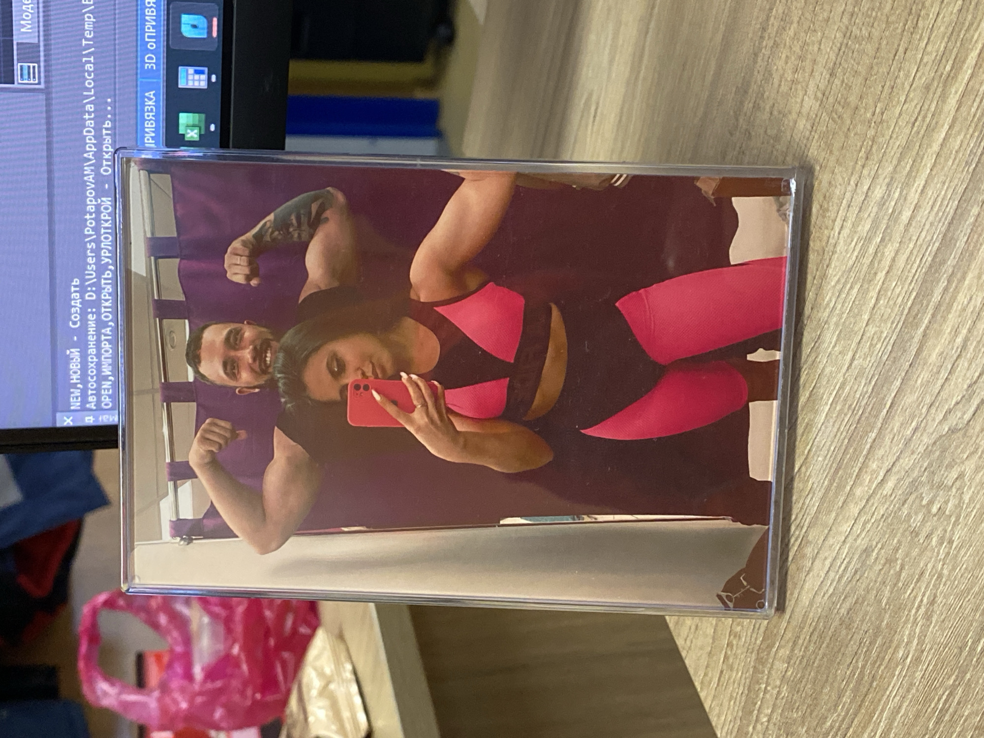 The image shows a photograph of a couple taking a selfie in a mirror, seemingly showcasing their fitness. The man has a tattoo on his arm. The woman is holding a red phone. The photo is in a plastic frame, which is placed on a wooden surface. In the background, there's a computer screen with text in Russian, indicating a software application. Below the photo, there is a partially visible plastic bag. No information in the image indicates the location to be Luzhitsy, Russia.