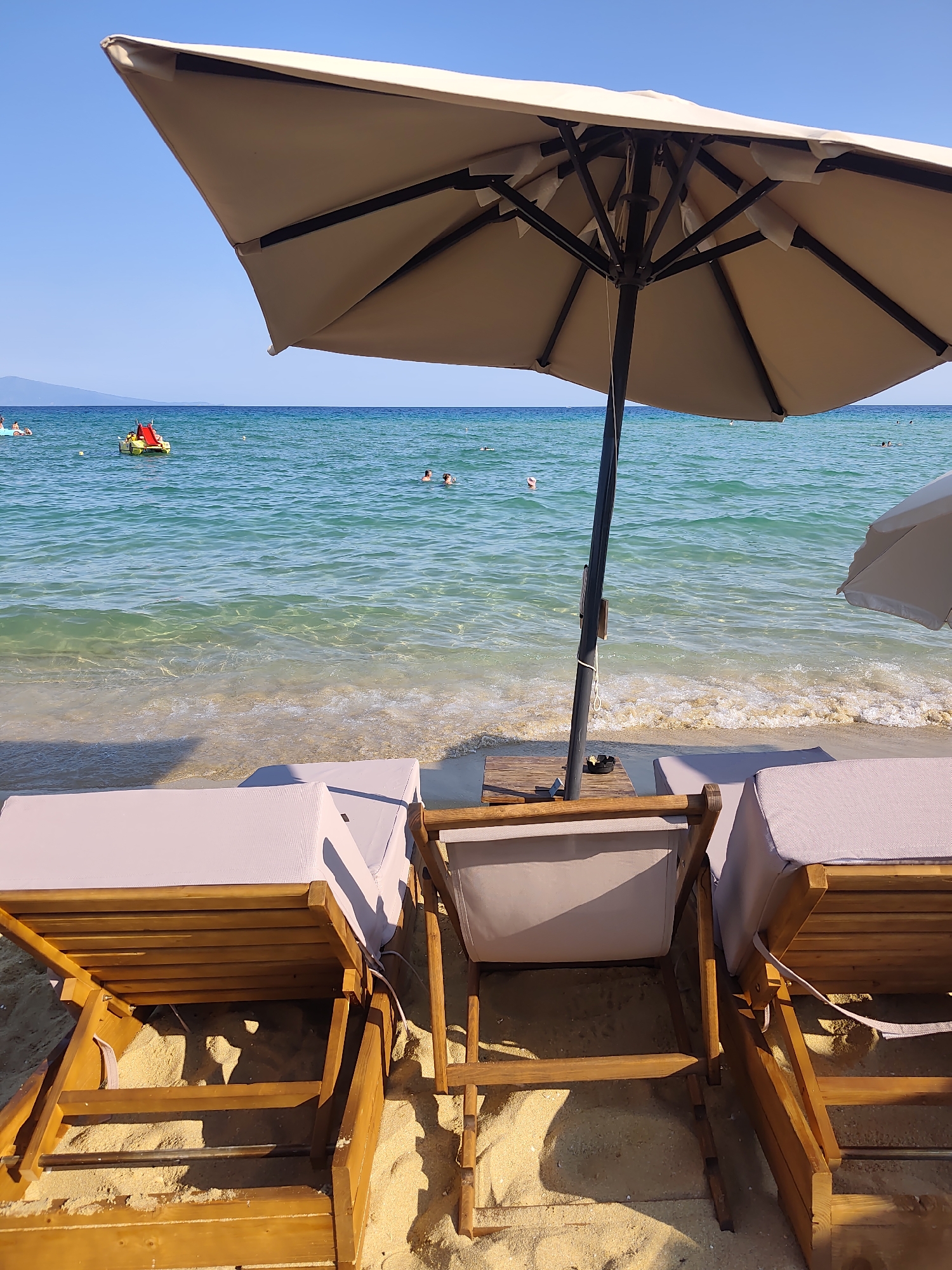 The image depicts a serene beach scene in Nea Peramos, Greece. In the foreground, two empty wooden lounge chairs with light-colored cushions are set up on the sandy beach, facing the turquoise waters of the Aegean Sea. A large, beige beach umbrella is open and positioned over the chairs, providing shade. To the right of the umbrella, a small wooden table holds an ashtray with cigarette butts. In the middle ground, the clear, shallow water gently laps the shore, with small waves breaking. Several people are visible in the sea, some swimming and others relaxing on inflatable toys. A yellow inflatable with a red slide is further out in the water, and a buoy is also visible. The background shows a clear blue sky and a distant coastline, possibly Mount Pangeo. The overall atmosphere is one of a peaceful summer day at the beach.