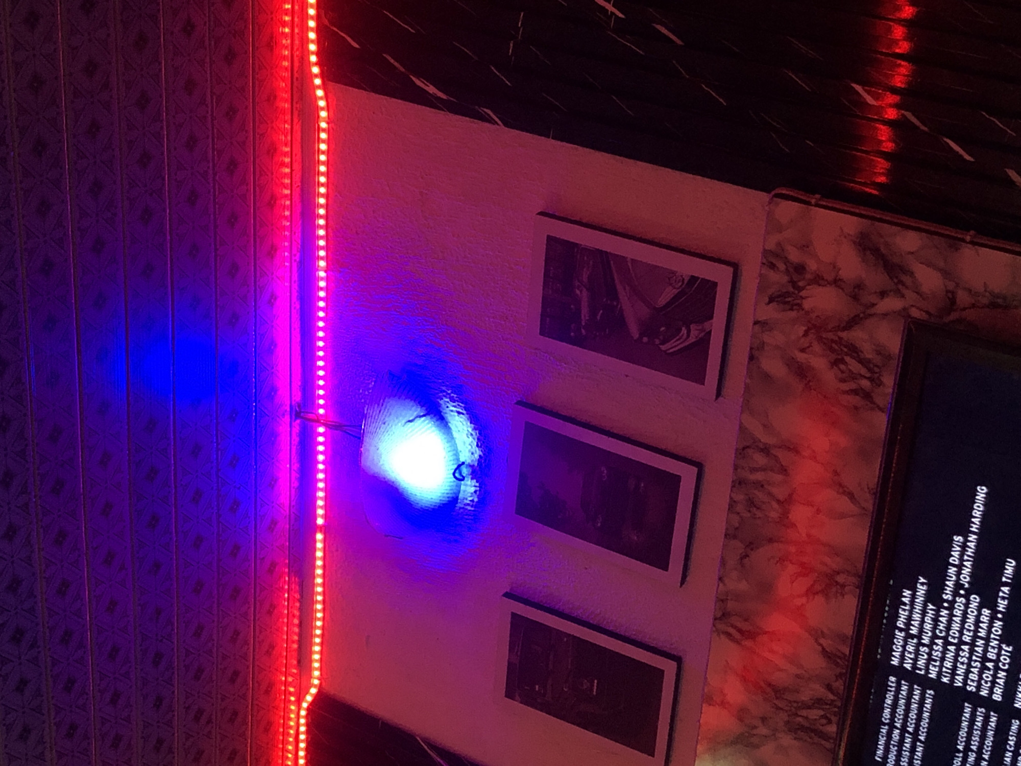 The image shows a wall with three framed pictures and an LED light strip. The light strip emits red light at the top and blue light below, creating a colorful ambiance. The framed pictures appear to be abstract art or possibly photographs. In the lower part of the image, a dark screen displays text, which seems to be a list of names and job titles, possibly credits from a film or production. The wall has a textured surface, with some parts appearing to be marble-patterned. The overall impression is that of an entertainment venue or a place decorated with a modern, artistic theme, possibly in New Stadium, Nigeria. There are no people visible in the image.