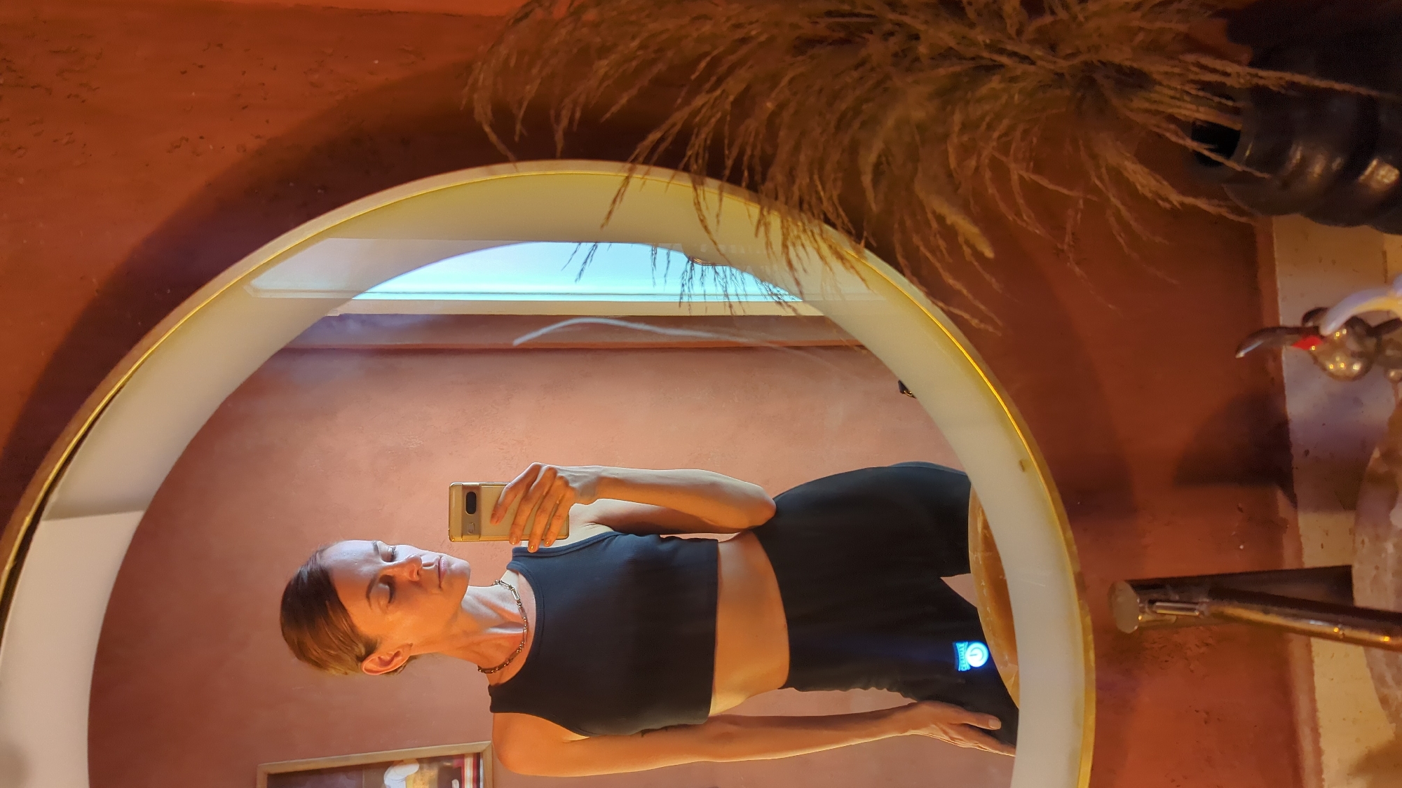 A woman is captured in a mirror selfie, her reflection dominating the frame. She is wearing a black crop top and black shorts, holding a light-colored smartphone (possibly a Google Pixel) in her left hand to take the picture. Her eyes are closed or looking downwards, and she has a neutral expression. A thin, golden-colored chain necklace is visible around her neck.
The mirror is uniquely designed with an arched shape and features an integrated light strip along its inner rim, emitting a soft glow. Surrounding the mirror is a textured wall, painted in a warm, reddish-brown hue. Above the mirror, dried pampas grass or similar ornamental reeds extend from a dark vase, creating a decorative element. To the right, a metallic fixture or decorative object is partially visible.
In the mirror's reflection, above the woman's head, a horizontal light source or window emits a bright, light blue light. Below the mirror, a framed picture is partially visible in the reflection. A small blue light or screen is visible on the side of the woman's left leg, possibly from a fitness tracker or a device in her pocket. The overall aesthetic, characterized by the textured wall and dried foliage, suggests a warm, possibly rustic or bohemian interior design, consistent with the style often found in places like North Kuta, Indonesia. No clear text is visible except for a partial, multi-colored logo (likely Google's) on the back of the smartphone. The image is presented on its side, as if taken in portrait orientation and then rotated.