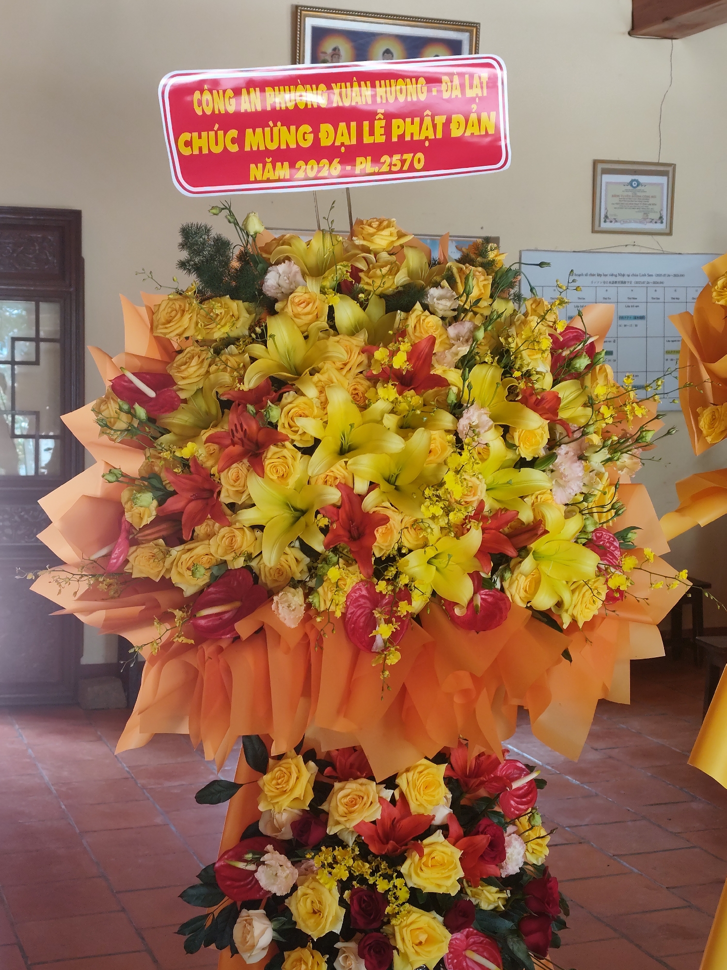 The image shows a large floral arrangement in Da Lat, Vietnam. Prominently displayed atop the bouquet is a sign reading "CÔNG AN PHƯỜNG XUÂN HƯƠNG - ĐÀ LẠT CHÚC MỪNG ĐẠI LỄ PHẬT ĐẢN NĂM 2026-PL.2570" indicating that the arrangement is from the Xuan Huong Ward Police Department in Da Lat, and it is celebrating Buddha's Birthday in 2026 (Buddhist calendar year 2570). The arrangement is primarily composed of yellow and red lilies and roses wrapped in orange tissue paper. In the background, there is a wooden cabinet and a framed picture of what appears to be a Buddha. There is also a whiteboard with what appears to be a class schedule.