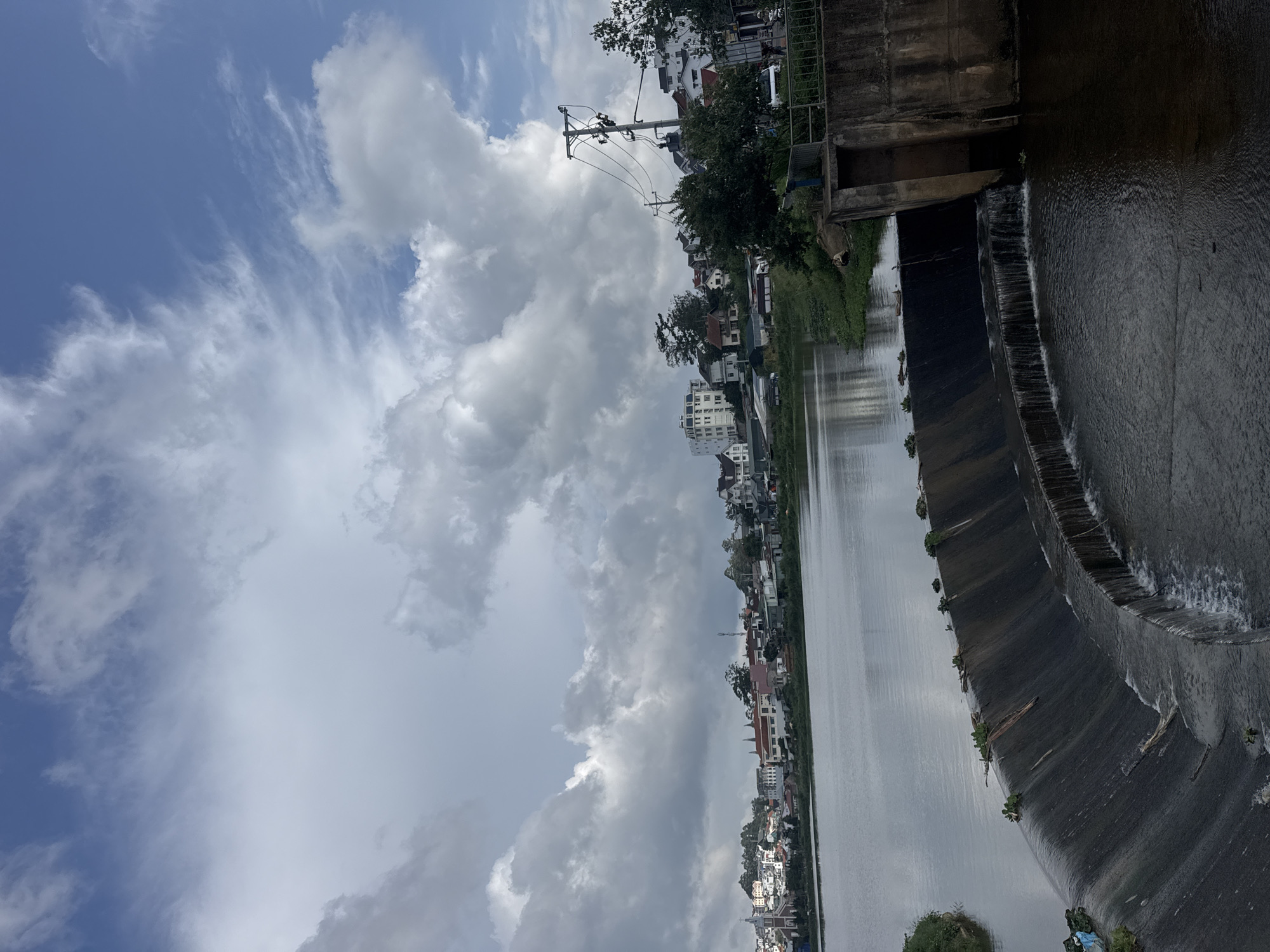 The image presents a wide view of a scenic urban landscape in Lâm Viên - Đà Lạt, Vietnam, featuring a substantial body of water alongside a town under a dynamic sky. The upper portion of the frame is dominated by a sky with a striking mix of clear blue patches and large, billowy white and grey cumulus clouds, interspersed with some wispy, high-altitude clouds.
A calm, expansive body of water, likely a lake or wide river, occupies the middle ground, reflecting the cloudy sky. Spanning the width of this water body is a concrete dam or weir, over which water cascades in several sections, creating small, uniform waterfalls and ripples on the downstream side. Lush green vegetation, including shrubs and trees, lines the banks of the water and grows on the dam structure itself.
Along one side of the water, a bustling residential area is visible, characterized by a continuous line of buildings of varying sizes and architectural styles, including houses and multi-story structures. Trees are dotted throughout the settlement, adding greenery to the urban environment. A prominent utility pole with multiple power lines stands out against the sky and buildings on the left bank.
The scene depicts daytime, likely late morning or early afternoon, given the bright illumination and visible blue sky. The weather appears to be partly cloudy, suggesting a blend of sunshine and cloud cover. No specific human activity is distinctly visible, and there is no discernible text within the image. The overall impression is one of a tranquil yet developed area by the water.
