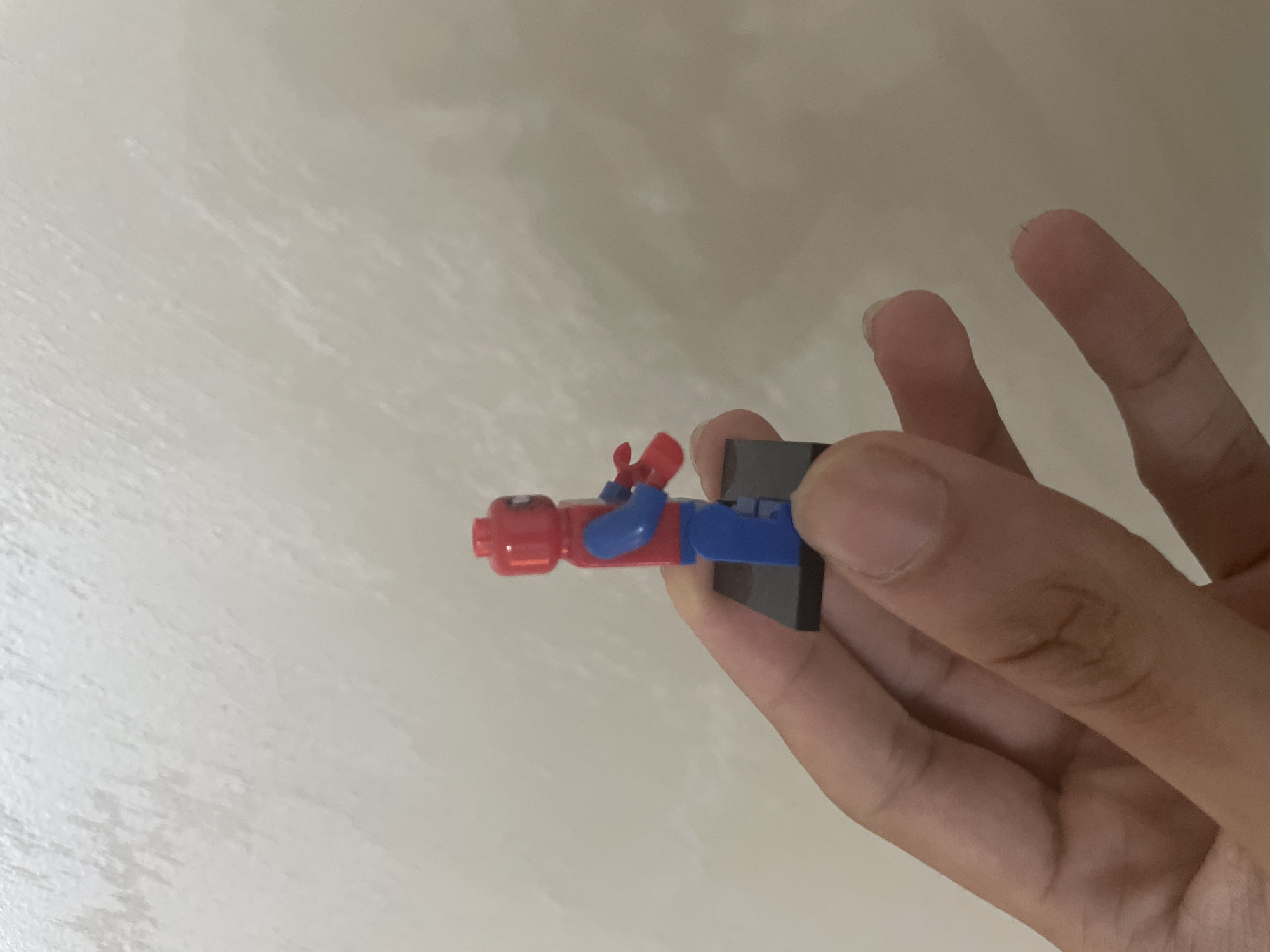 The scene depicts a human hand holding a Lego minifigure against a plain, textured wall, occurring in the city of Ihtiman, Bulgaria. The hand, with light to medium skin tone, occupies the lower portion of the frame, with visible fingers and knuckles. The Lego minifigure is predominantly red and blue, featuring a red head with indistinct facial markings, a red torso with blue arms, and blue legs. The minifigure's left arm is bent, and its hand holds a small, red, indistinct accessory. It stands on a rectangular black Lego display base (2x3 studs). The background is a uniformly lit, light beige or off-white wall with subtle, irregular surface texture.