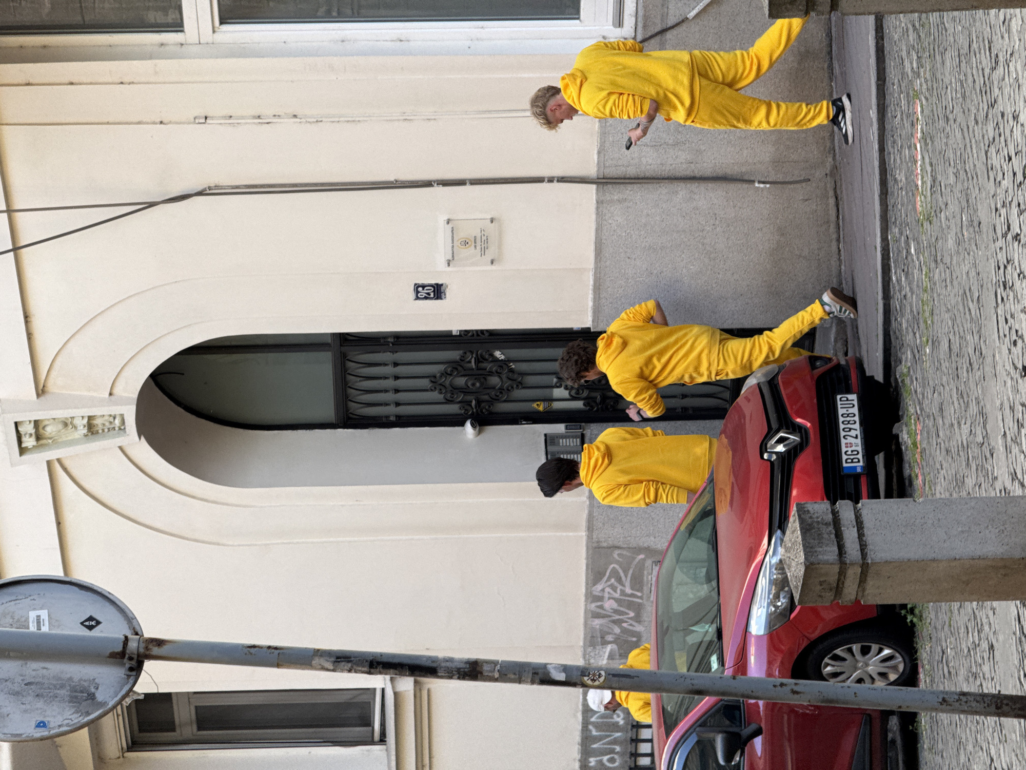The image shows an exterior street scene in Belgrade, Serbia. The foreground contains a weathered gray pole with a circular road sign. A red Renault car is parked on the right side of the frame. Three people wearing bright yellow tracksuits are positioned alongside the car and against a light-colored building with an arched entrance. The building features a black ornate metal gate and a number plaque "96". The license plate of the vehicle reads "BG 2988-UP". The architecture, the license plate and the sign suggests that the location is likely in Belgrade, Serbia.