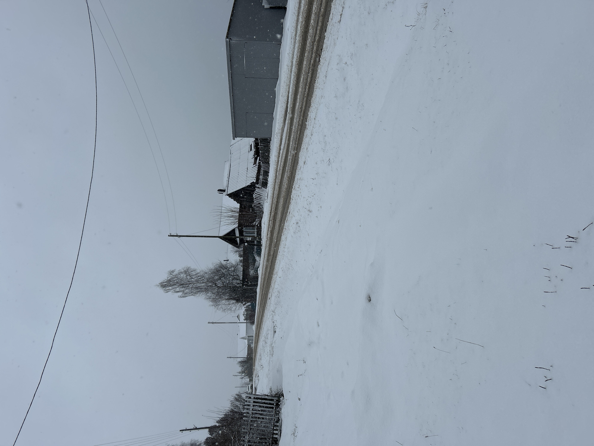 The image depicts a snow-covered scene in Уральское, Russia. A road runs through the middle, with tire tracks visible in the snow. To the left of the road are houses, trees, and telephone poles with lines. One of the buildings appears to be a large, gray structure. The sky is overcast and gray. The foreground is covered in snow with scattered debris.