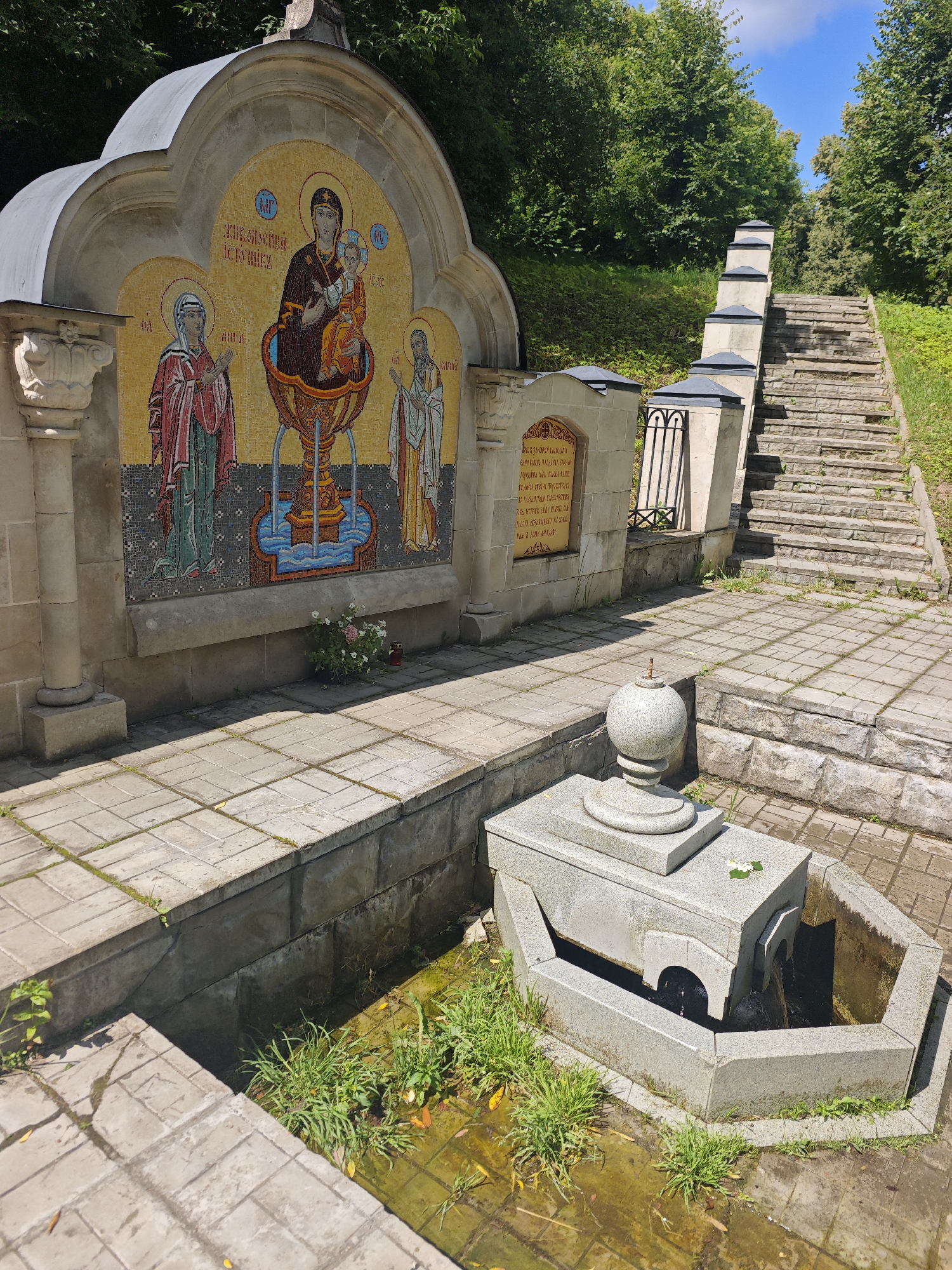 The image depicts a holy spring dedicated to Saint Nicholas in the city of Chekhov, Russia. The spring is located outdoors, with a backdrop of lush green trees and a blue sky with scattered clouds. In the foreground, there is a stone structure with a basin where water flows. The water appears somewhat murky, with visible green algae and a few fallen leaves. Surrounding the basin is a paved area made of light-colored stone tiles.
To the left of the spring is a large, arched mosaic depicting religious figures, likely Saint Nicholas, the Virgin Mary, and the infant Jesus, set against a golden background. The mosaic is integrated into a stone wall adorned with classical columns. In front of this mosaic, a small bouquet of pink and white flowers and a red candle are placed on the ground.
Adjacent to the mosaic structure and extending upwards is a stone staircase with a protective railing. The stairs lead up a grassy slope, further emphasizing the natural setting of the location. A stone plaque with text is affixed to the wall to the right of the mosaic, but the inscription is not clearly legible. The overall scene suggests a place of religious significance and natural beauty.