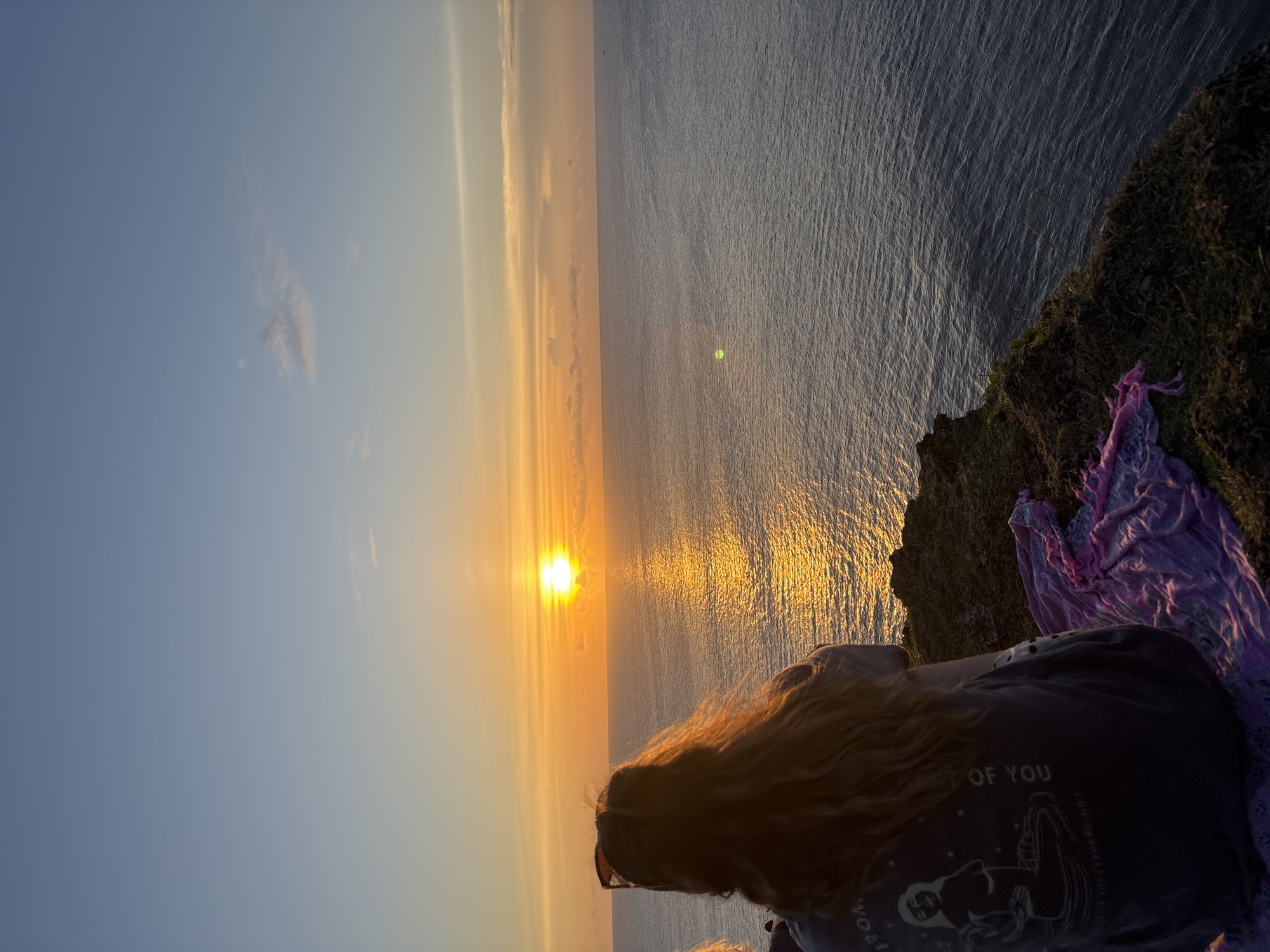 A woman with long blonde hair is standing on a cliff overlooking the ocean at sunset in Pecatu, Indonesia. The sun is setting over the water, creating a golden reflection on the surface. The cliff is covered in grass and there is a purple cloth draped over the edge. The woman is wearing a dark t-shirt with a graphic on it.