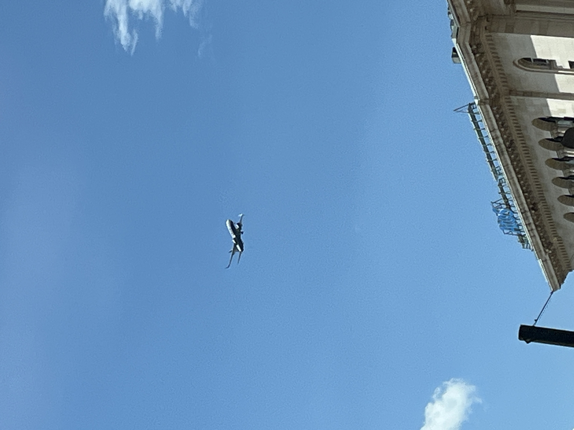 An airplane is flying in the sky above a building. The airplane is white with a black underbelly and is angled slightly downwards. It is in the upper middle portion of the image. Below the airplane, a beige-colored building with an ornate facade and arched windows is visible. A dark pole is in the lower left of the image. The sky is clear and blue with a few wispy white clouds.