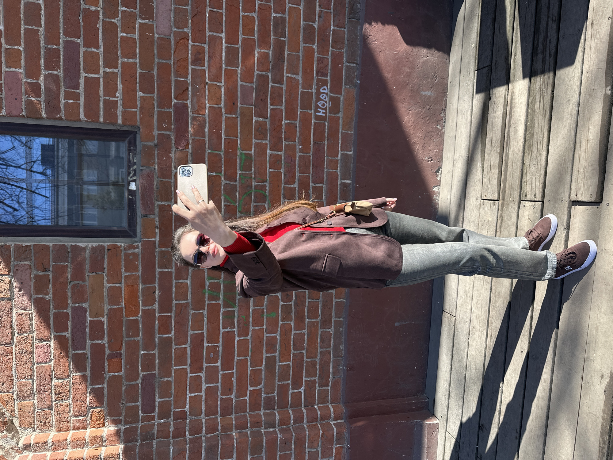A young woman is taking a selfie in front of a red brick building. She is wearing sunglasses, a red turtleneck sweater, a brown jacket, gray jeans, and brown sneakers. She is holding a tan-colored smartphone. The building has a window on the left side. The word "HOOD" is written in white on the building's brick, above the woman. The woman is standing on a wooden surface. The location is Kazan, Russia.