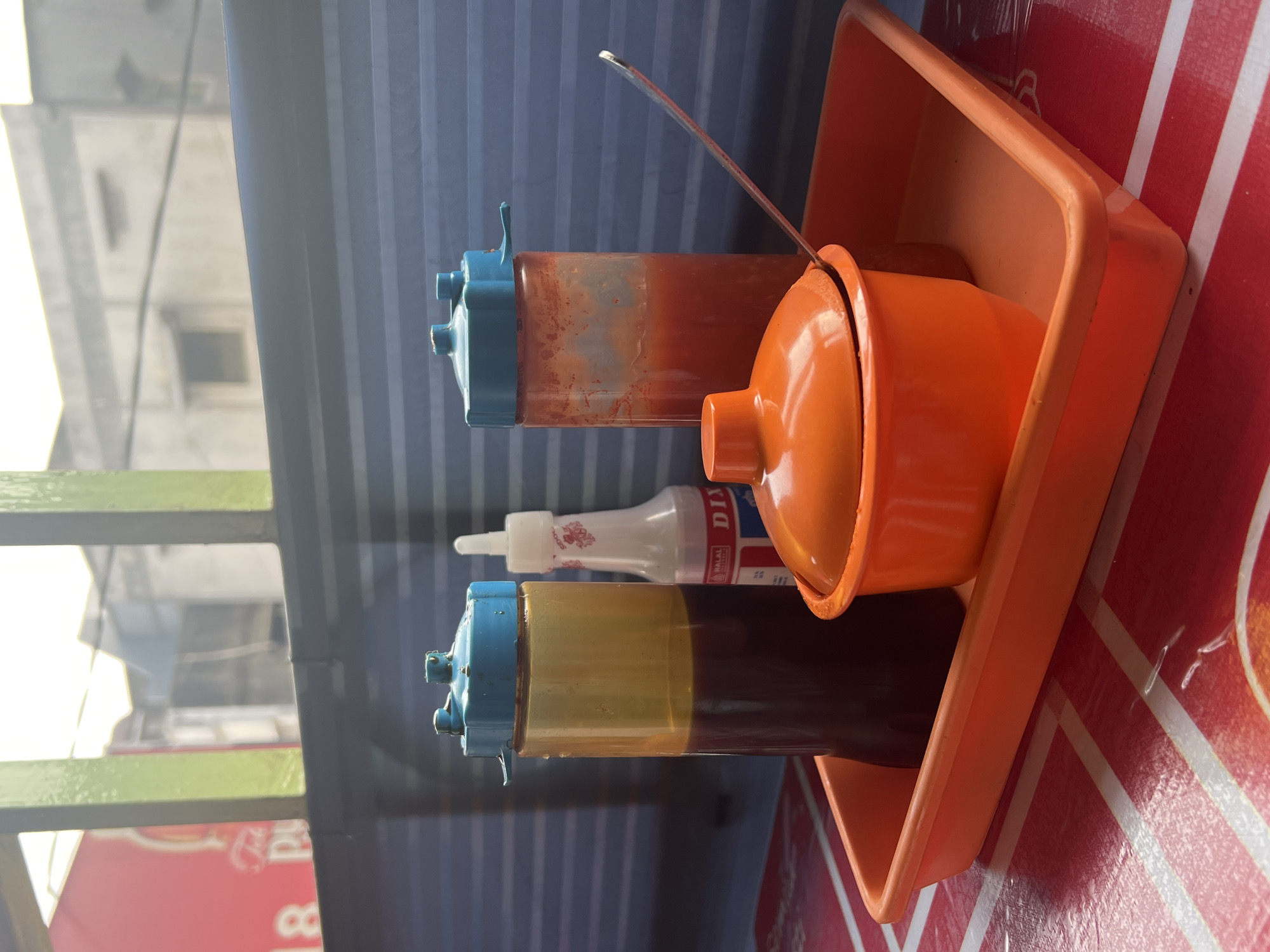 On a table with a red and white striped plastic cover, an orange rectangular tray holds three containers of condiments. On the left is a tall, translucent yellow bottle with a blue cap, filled with a dark brown liquid, likely soy sauce. To its right is a smaller white bottle with a red logo and white cap, with text that appears to say "DIYAL." In the center of the tray is an orange lidded bowl. To the right of the bowl is a tall, translucent red bottle with a blue cap, filled with a red liquid, likely chili sauce. A silver spoon rests on the edge of the tray, its handle extending towards the red condiment bottle. In the background, out of focus, is a blue corrugated wall, and beyond that, a window with green bars reveals buildings and a bright sky, suggesting an outdoor or semi-outdoor setting. The scene appears to be in a food stall or restaurant. Given the information that this is located in Bandung City, Indonesia, it is highly probable this is an outdoor dining area of a local eatery, common in Indonesian cities.