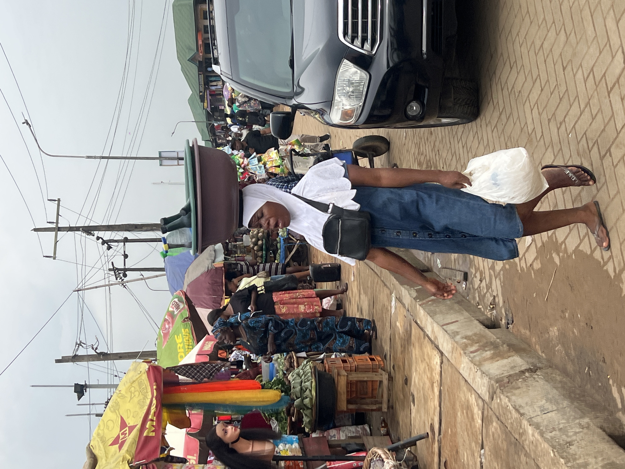 A market scene unfolds in Egan, Nigeria. A woman balances a large brown bowl on her head while walking along a brick-paved street. She wears a white headscarf, a white top, a denim skirt, sandals, and has a black bag slung across her body. She holds a white plastic bag in her right hand.
To her left, market stalls are covered with colorful umbrellas and display various goods. Other people are browsing or selling at the stalls. A mannequin head with long black hair is visible among the goods.
To her right, a black SUV is parked at the edge of the road. The street appears crowded. Power lines crisscross overhead against a cloudy sky. A concrete curb separates the street from a lower, dirtier area.